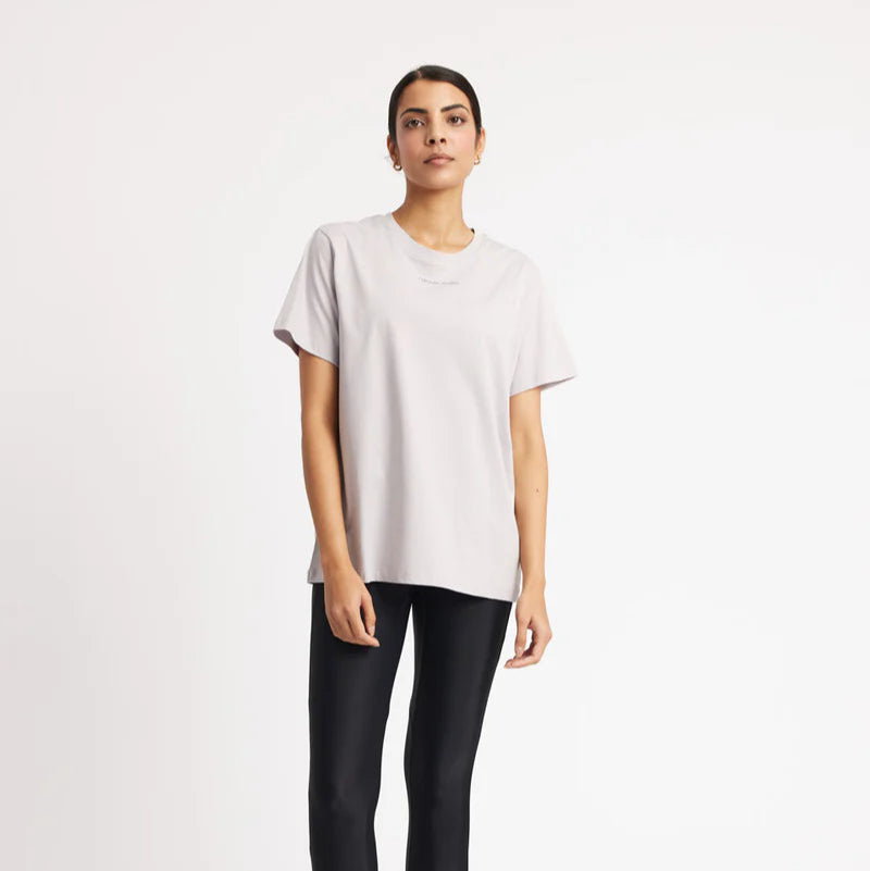 Eine Frau steht vor einem schlichten weißen Hintergrund und trägt ein lockeres hellgraues T-Shirt, schwarze Leggings und graue Sportschuhe. Sie blickt mit neutralem Gesichtsausdruck und entspannter Körperhaltung nach vorne.