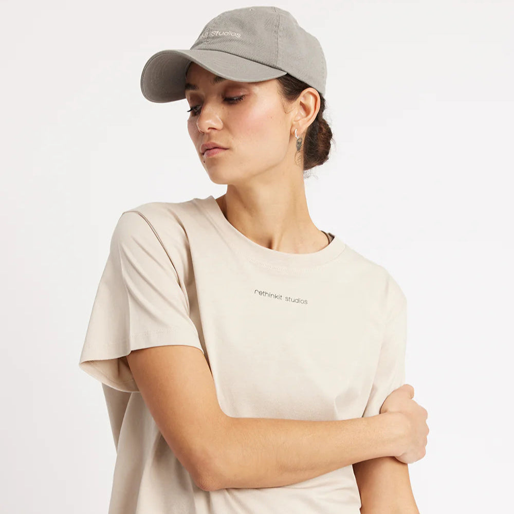Eine Frau, die ein beiges T-Shirt mit einem anderen Studiotext auf der Brust und eine hellgraue Baseballkappe trägt, steht vor einem schlichten weißen Hintergrund und blickt leicht nach unten, wobei sie einen Arm über dem Körper verschränkt.
