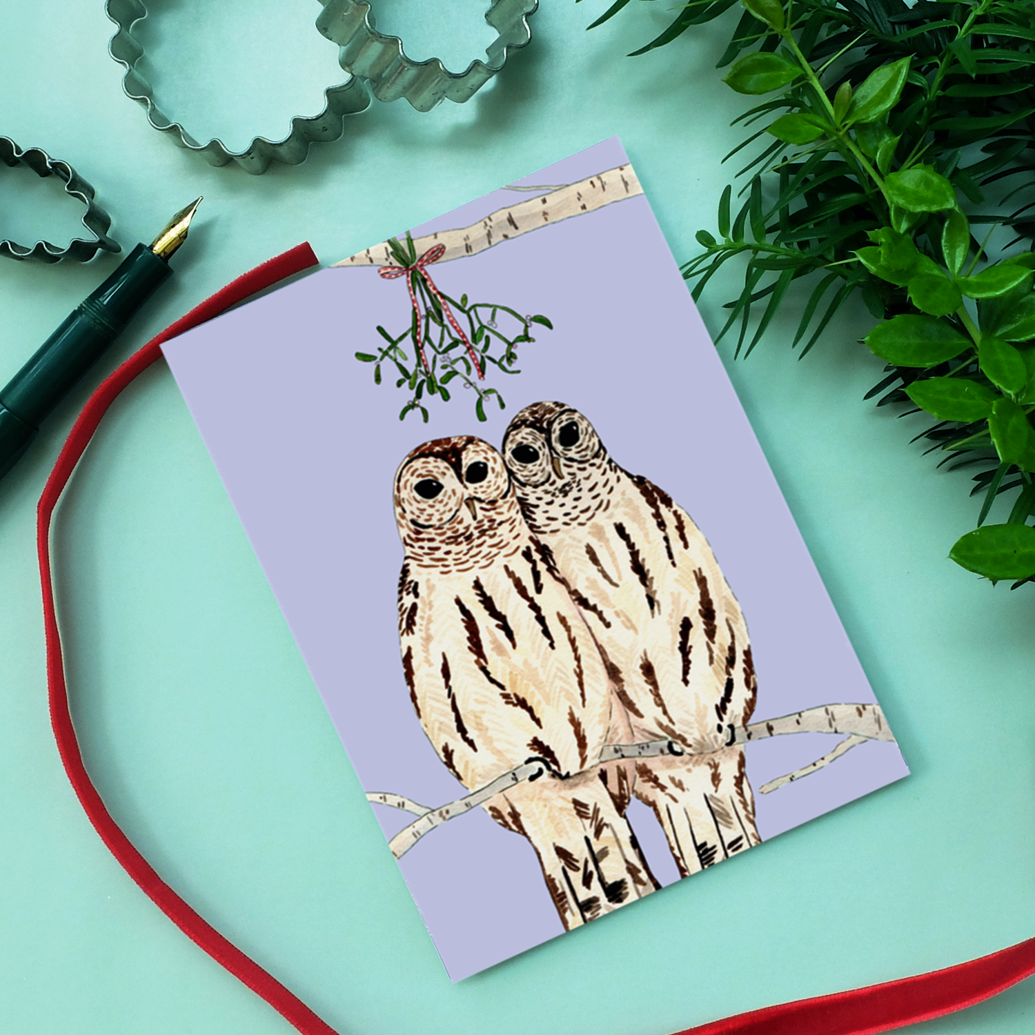 Eine Grußkarte mit zwei illustrierten Eulen, die nebeneinander auf einem Ast mit einem Mistelzweig über ihnen hocken. Die Karte ist umgeben von einem roten Band, einem grünen Stift, einer Pflanze und einem Ausstecher auf einer hellblauen Oberfläche.