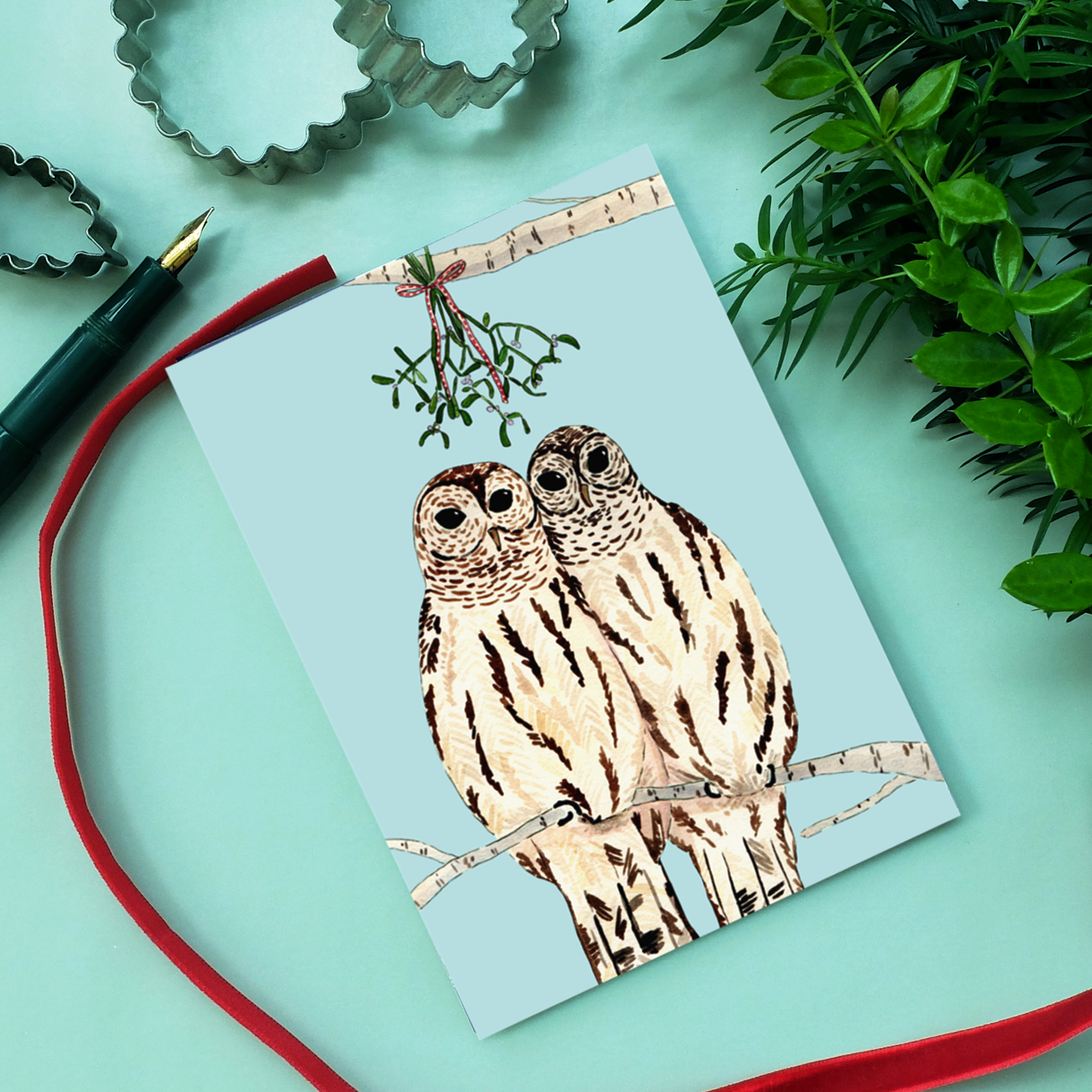 Eine Grußkarte mit zwei illustrierten Eulen auf einem Ast unter einem Mistelzweig, umgeben von einem roten Band, einem Füllfederhalter, Ausstechern und grünem Laub auf einer hellblauen Fläche.