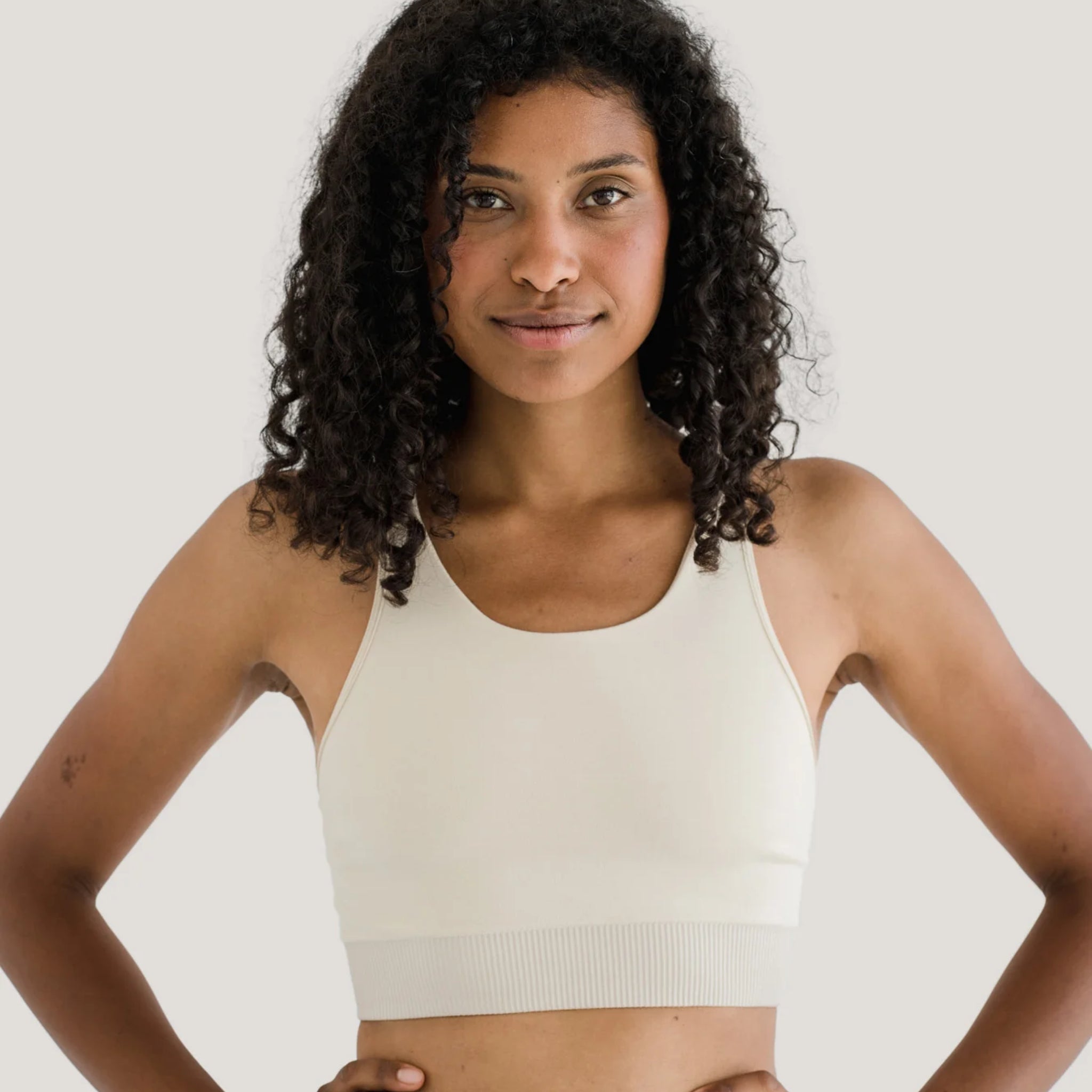 Frau mit lockigem Haar trägt ein cremefarbenes Sporttop und lächelt, vor einem neutralen Hintergrund.