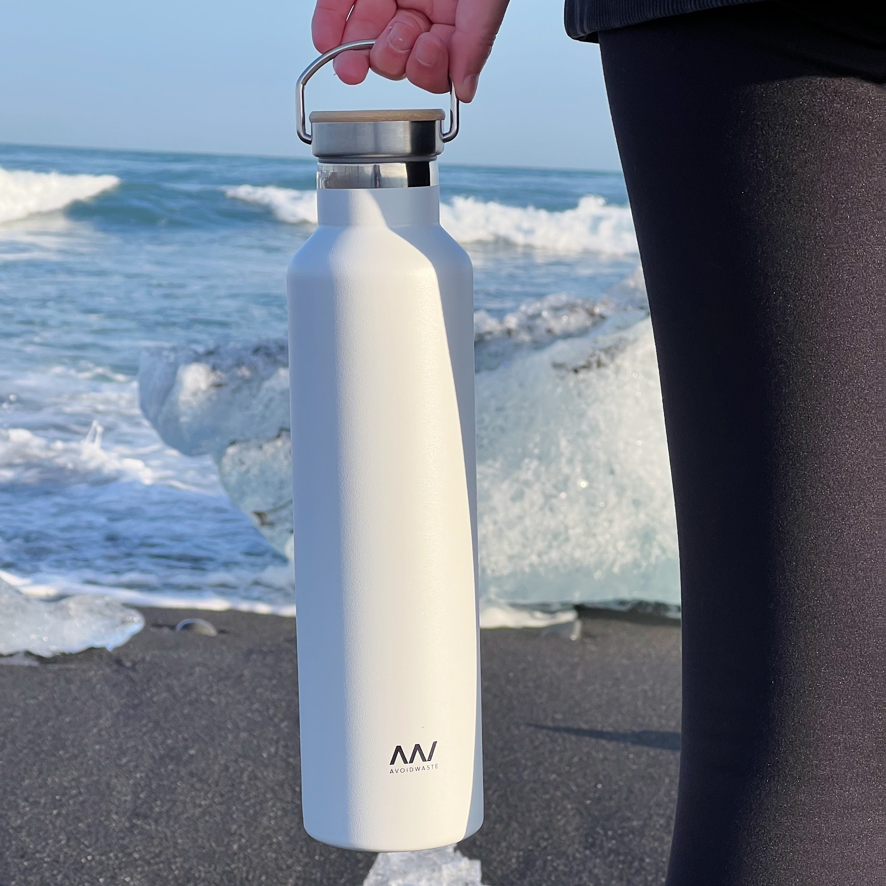 Eine Person hält eine weiße Trinkflasche am Strand, im Hintergrund das Meer und Eisbrocken.