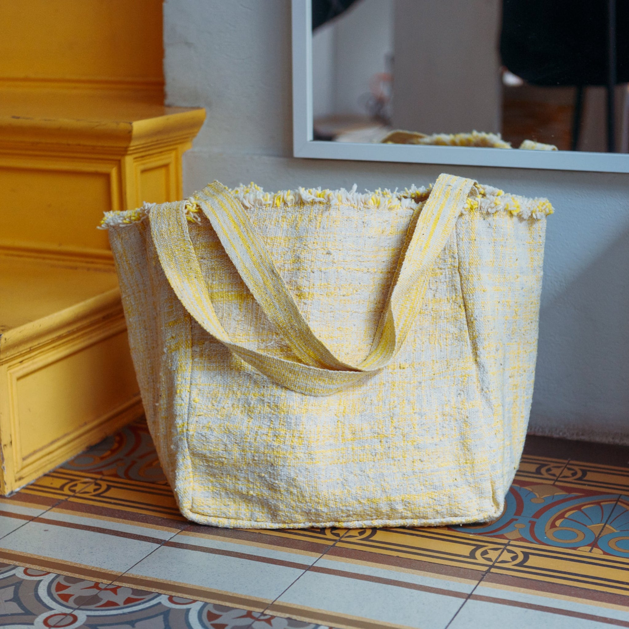 Gelbe Stofftasche mit Fransen steht auf gemustertem Boden neben gelber Stufe und Spiegel an der Wand.