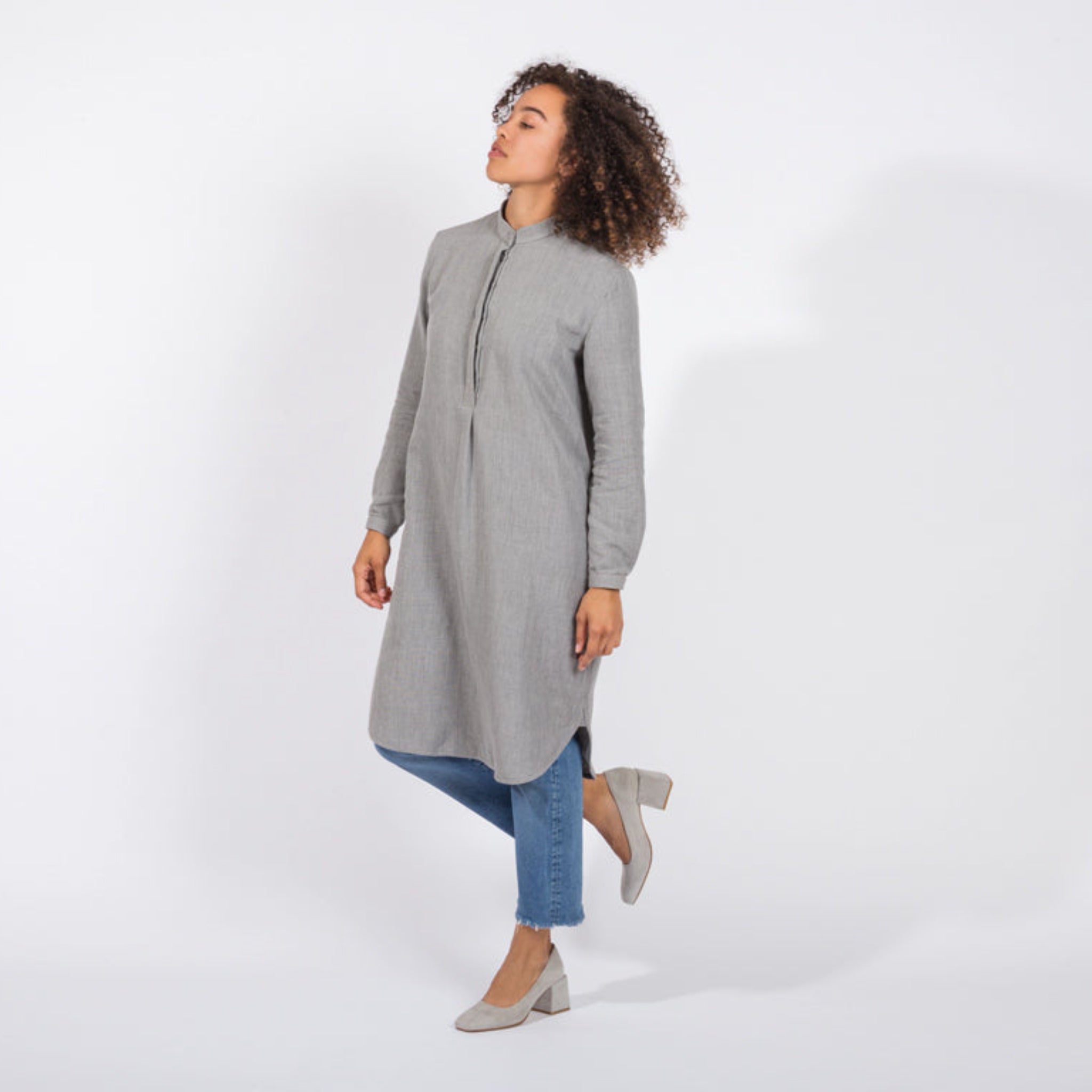 Person in grauem Kleid und blauen Jeans, steht mit einem Bein angehoben vor weißem Hintergrund.