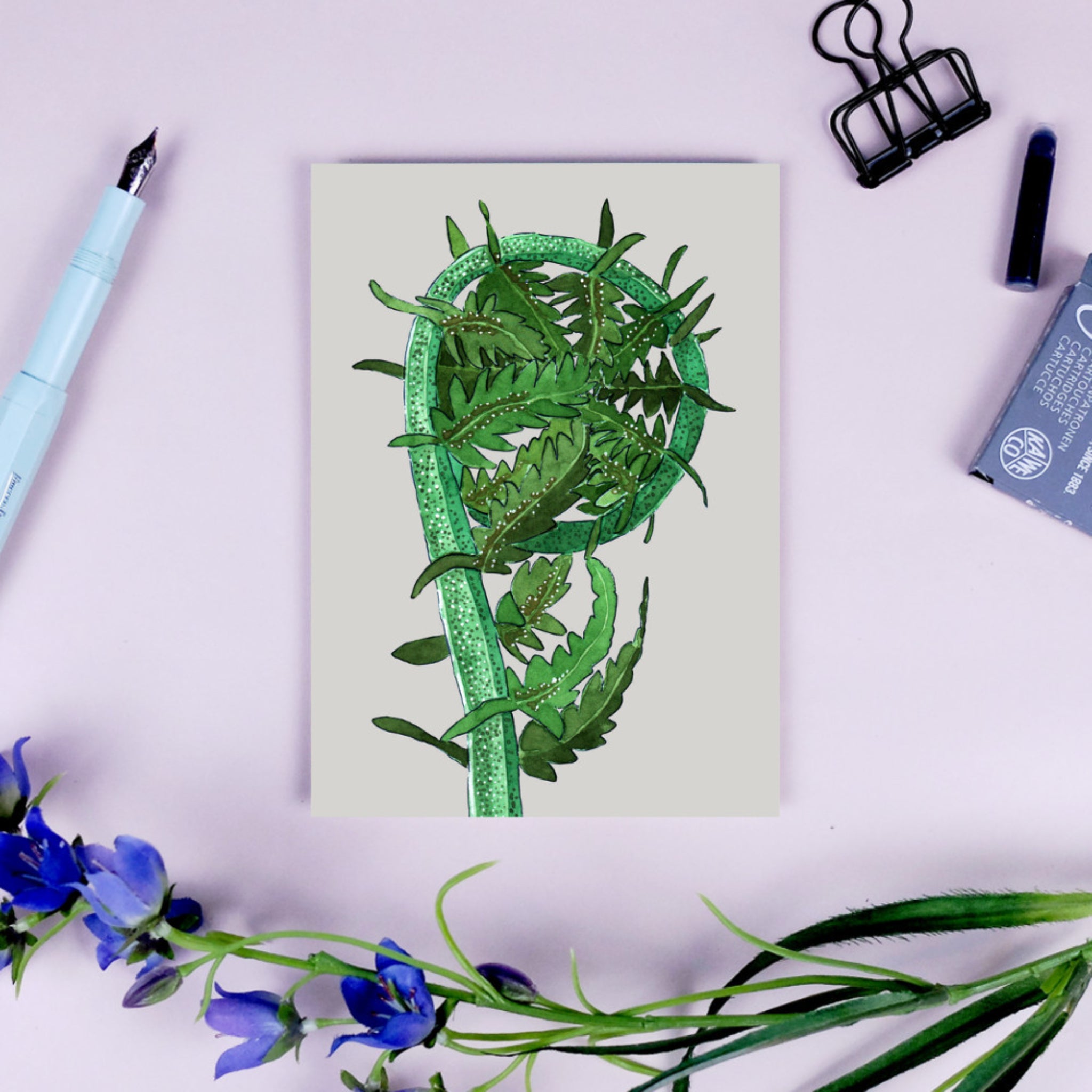 Postkarte mit grünem Farn auf einem Schreibtisch, umgeben von einem Stift, einer Büroklammer und blauen Blumen.
