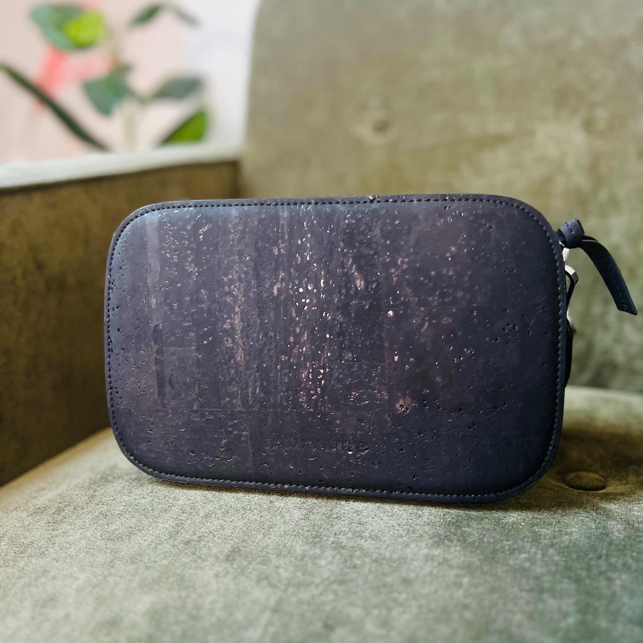 Eine schwarze Tasche aus strukturiertem Material liegt auf einem grünen Stoffsofa, im Hintergrund ist eine Pflanze.