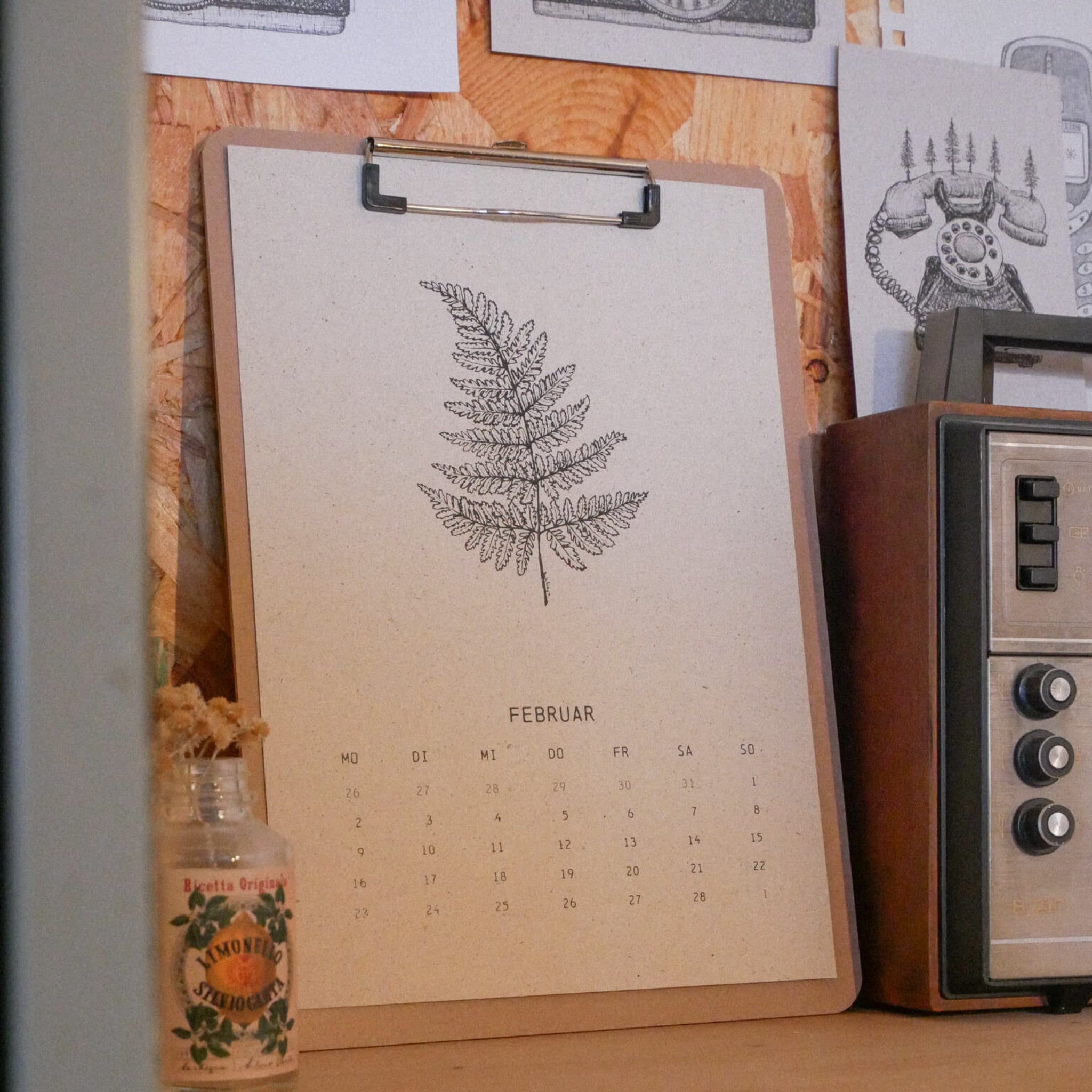 Ein Kalenderblatt für Februar mit einer detaillierten Abbildung eines Farns ist an ein Brett geklebt. Rechts steht ein altes Radio und links eine kleine Glasflasche mit der Aufschrift "Almond". An der Wand dahinter sind Schwarz-Weiß-Zeichnungen angeheftet.