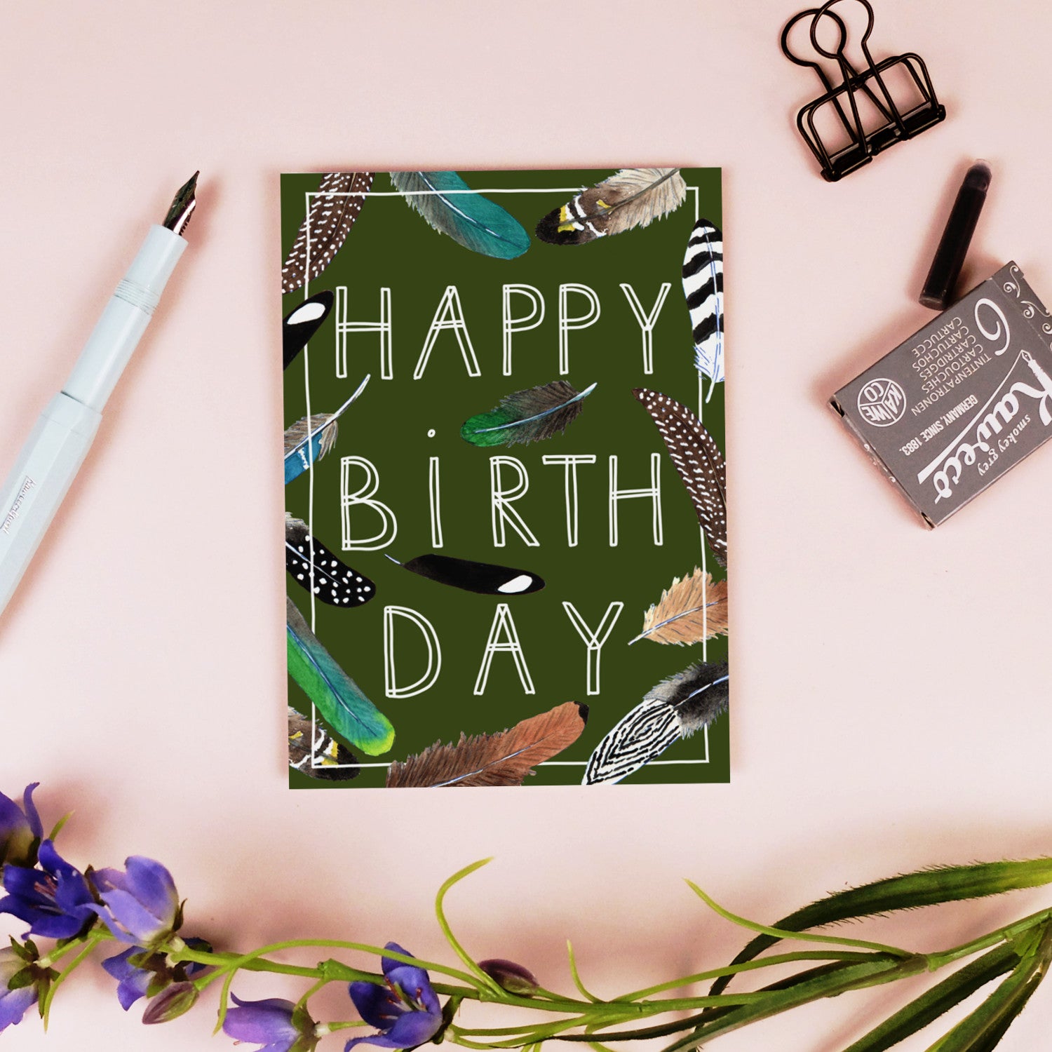 Grüne Geburtstagskarte mit Federn und "Happy Birthday"-Text, umgeben von einem Stift, einer Klemme und Blumen.