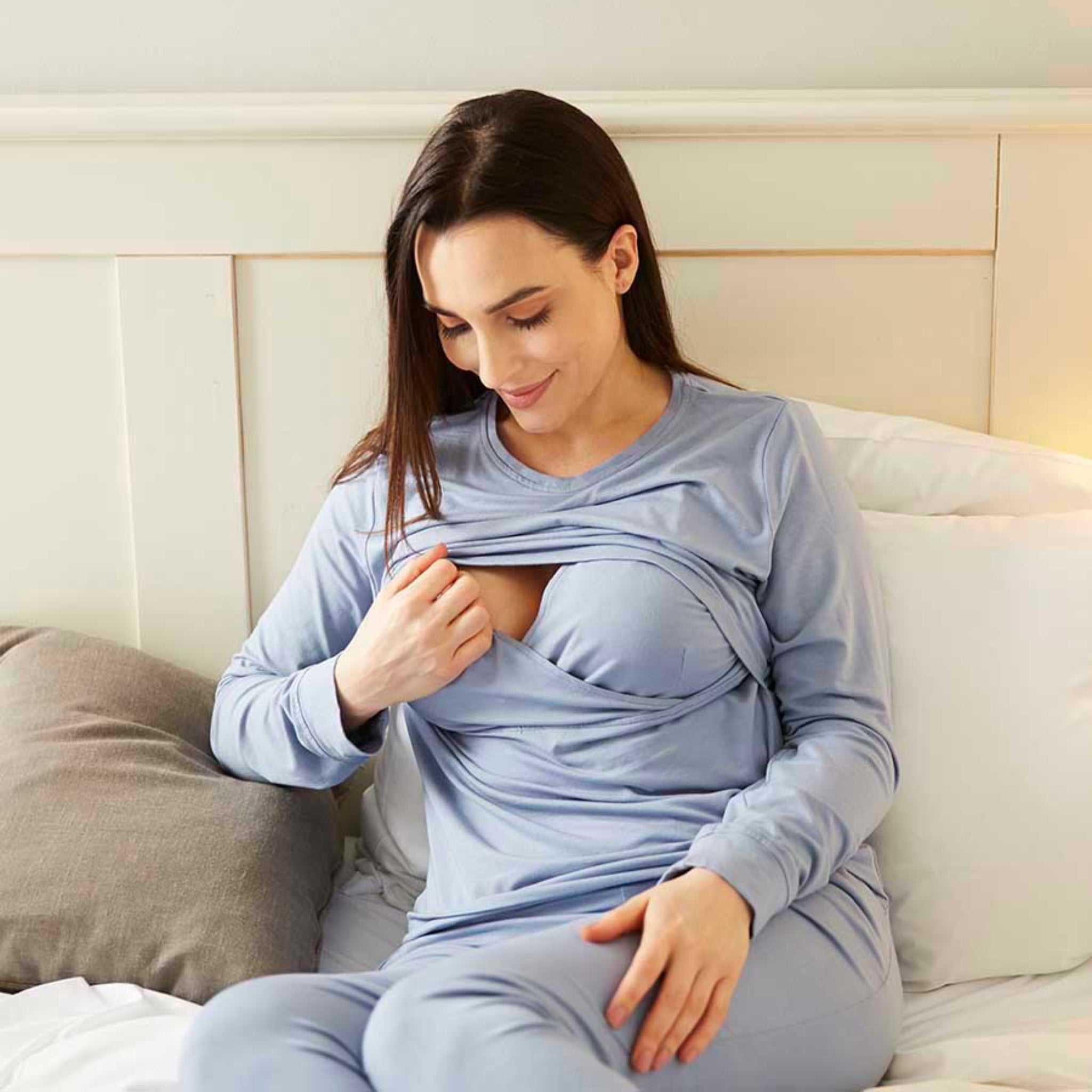 Eine Frau in blauer Stillkleidung sitzt auf einem Bett und hebt den Stoff, um das Stillen zu erleichtern.