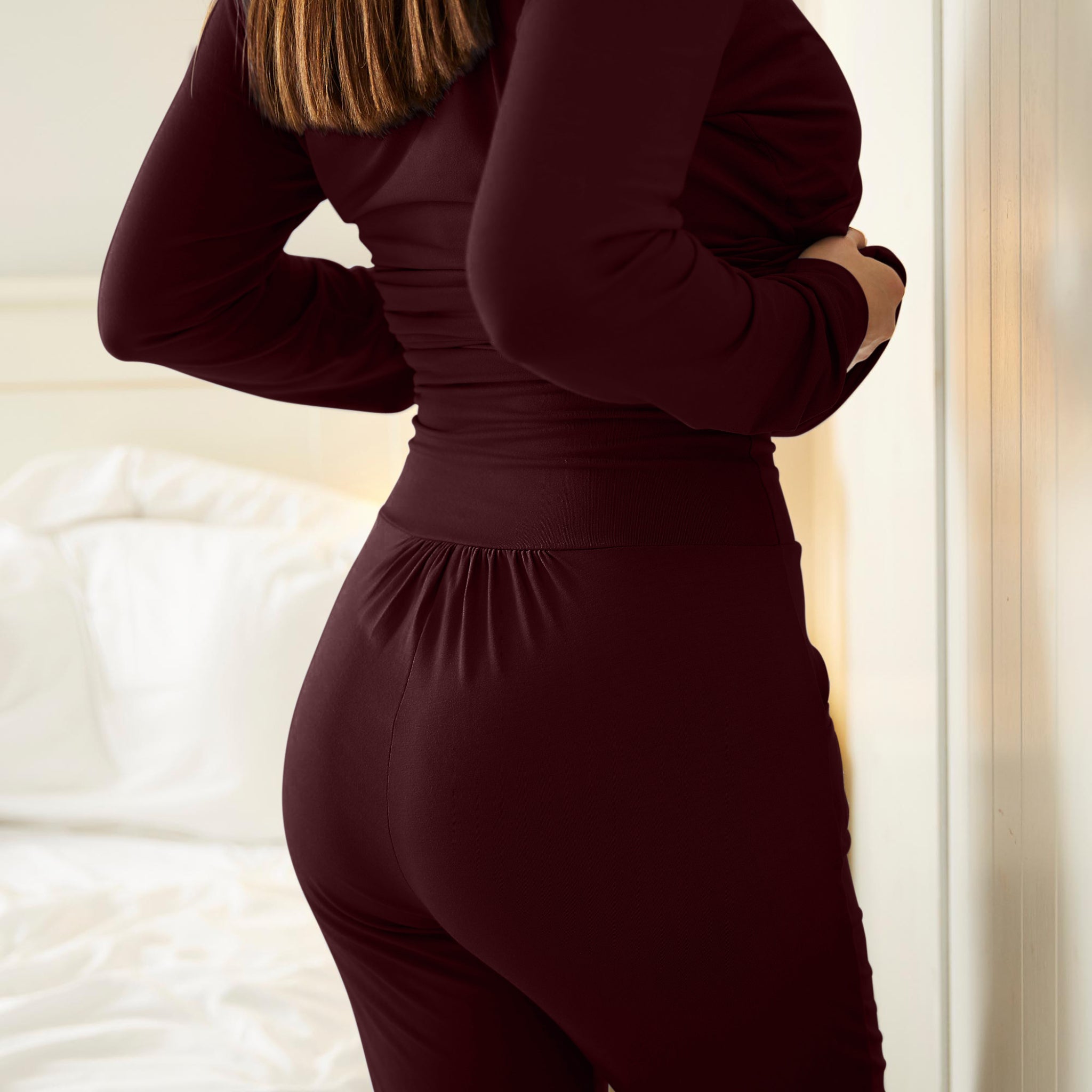 Person in dunkelrotem Outfit steht vor einem weißen Bett und hält sich am Oberkörper.