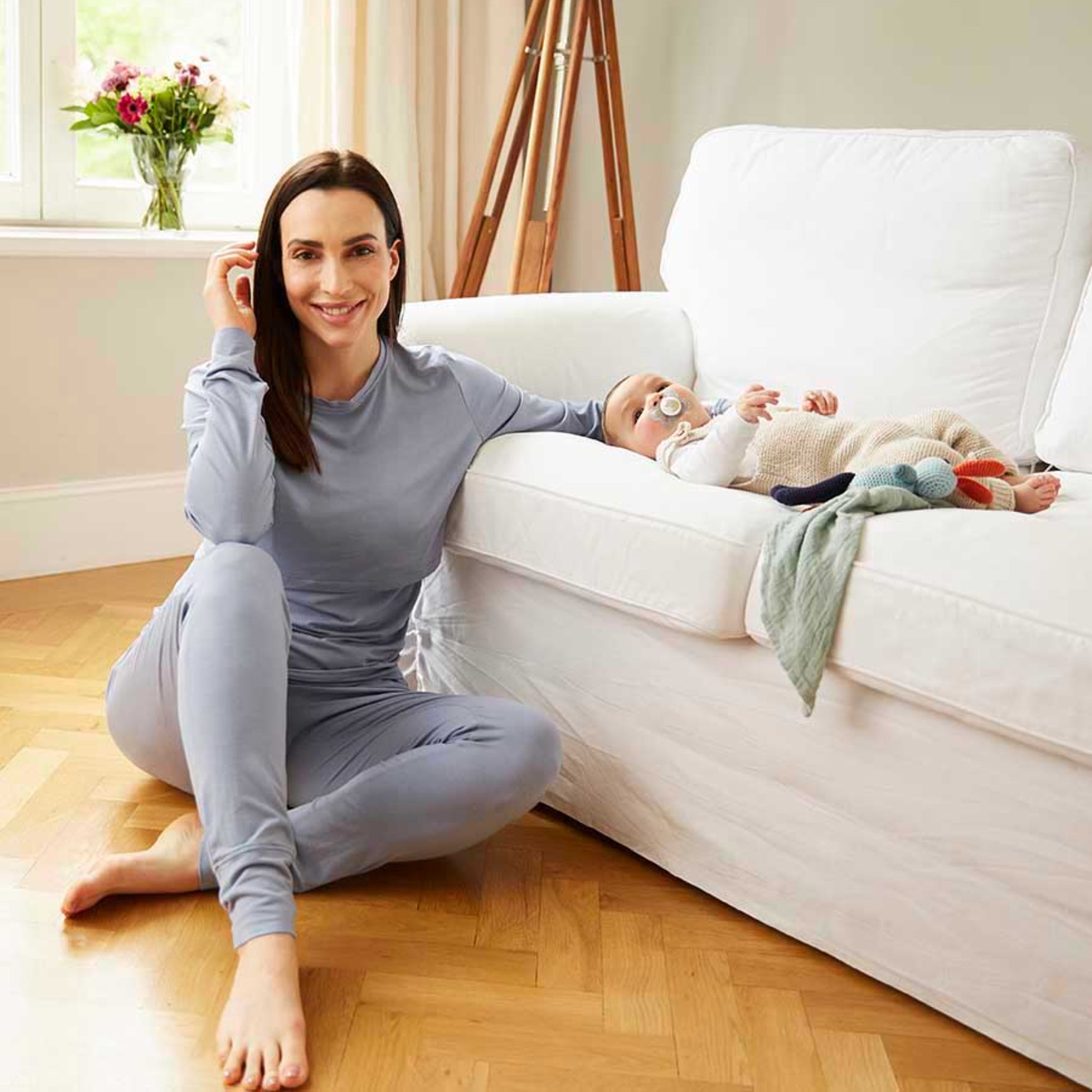 Frau in grauem Pyjama sitzt neben einem Sofa, auf dem ein Baby mit Schnuller und Kuscheltier liegt.