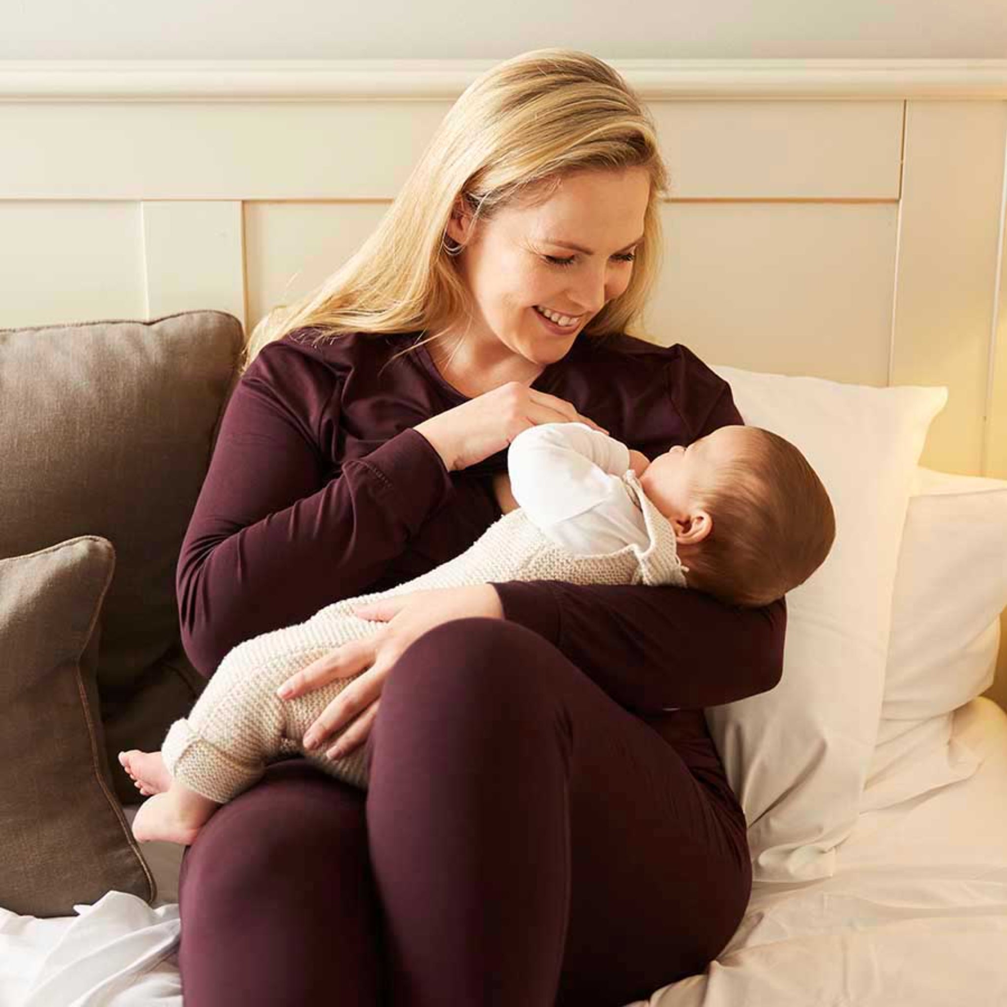 Frau sitzt lächelnd auf einem Bett und hält ein Baby in den Armen, umgeben von Kissen.