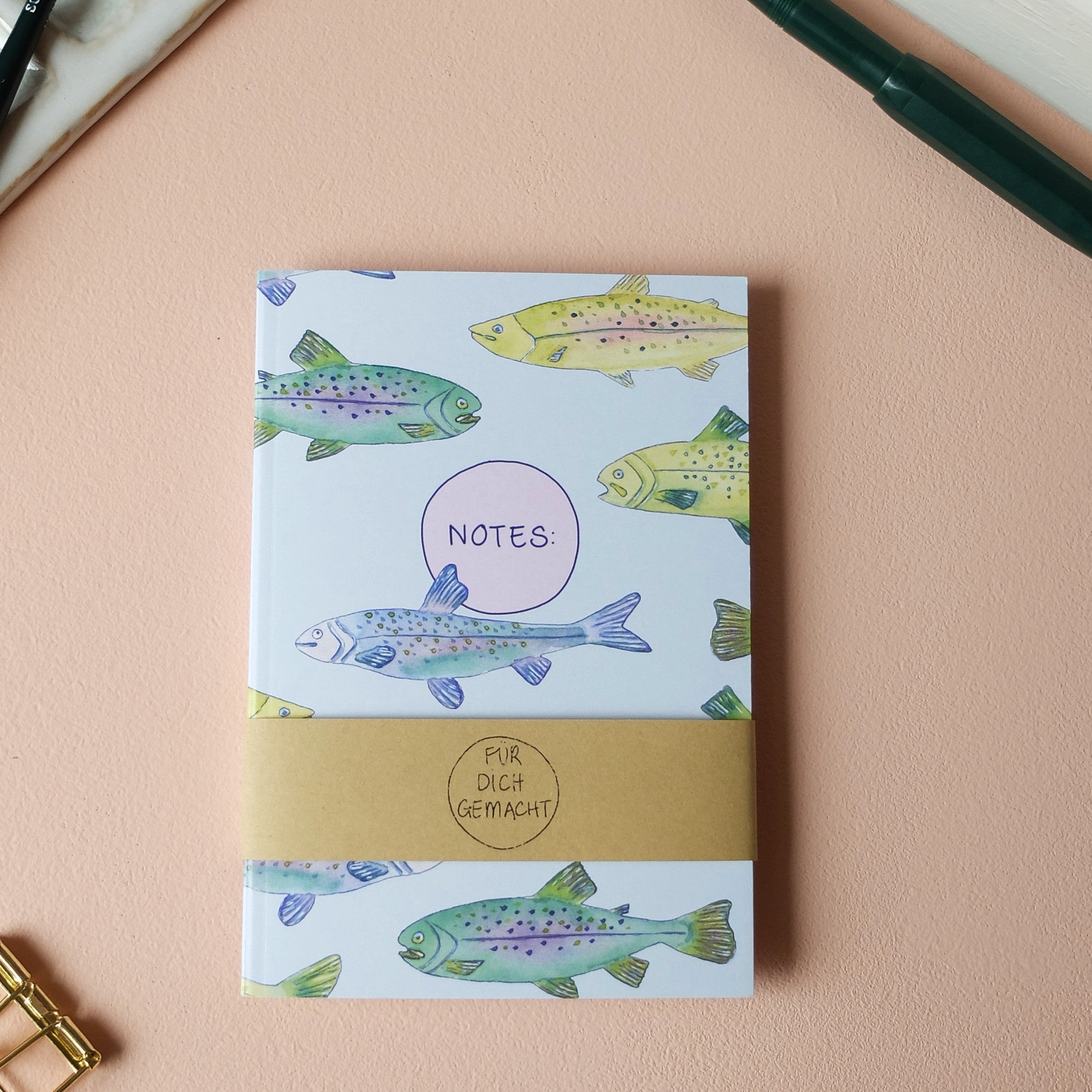 Ein Notizbuch mit bunten Fisch-Illustrationen auf dem Cover liegt auf einem Tisch mit einer Stift und einer Büroklammer.