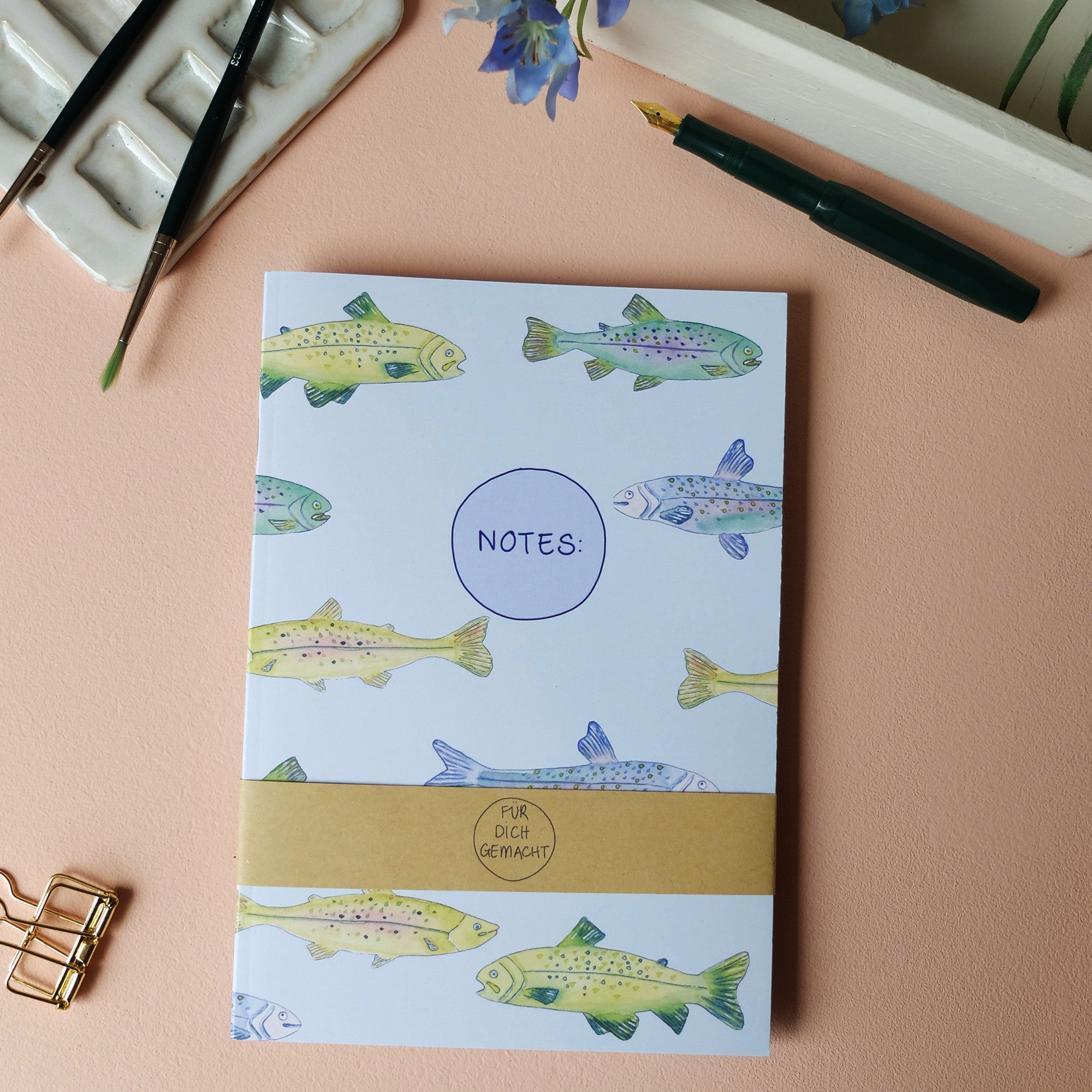 Notizbuch mit Fischmuster auf einem Tisch, mit Pinsel, Füller und lila Blumenarrangement daneben.
