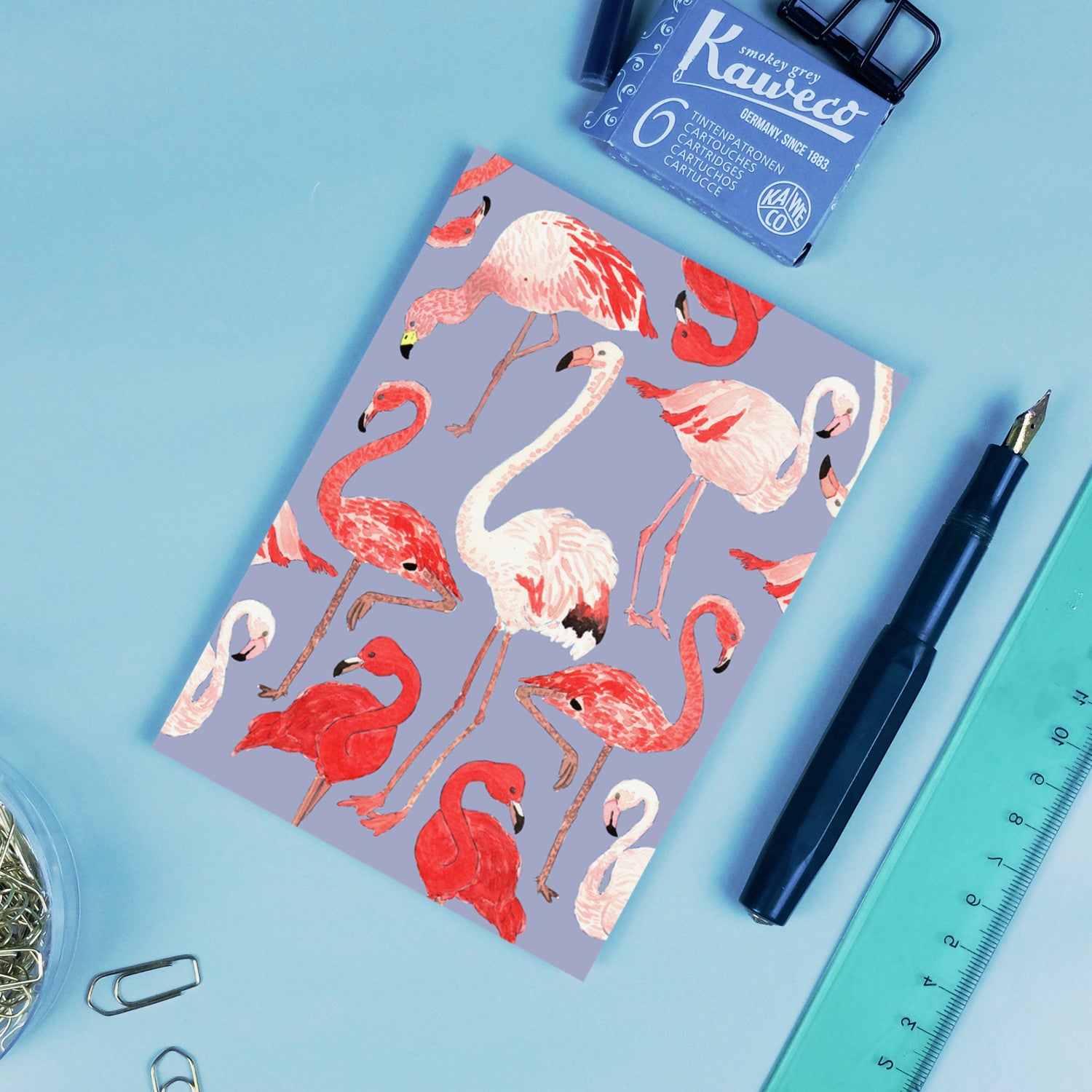 Notizbuch mit rosa und roten Flamingos auf blauem Hintergrund, umgeben von Stift, Lineal und Tintenpatronen.
