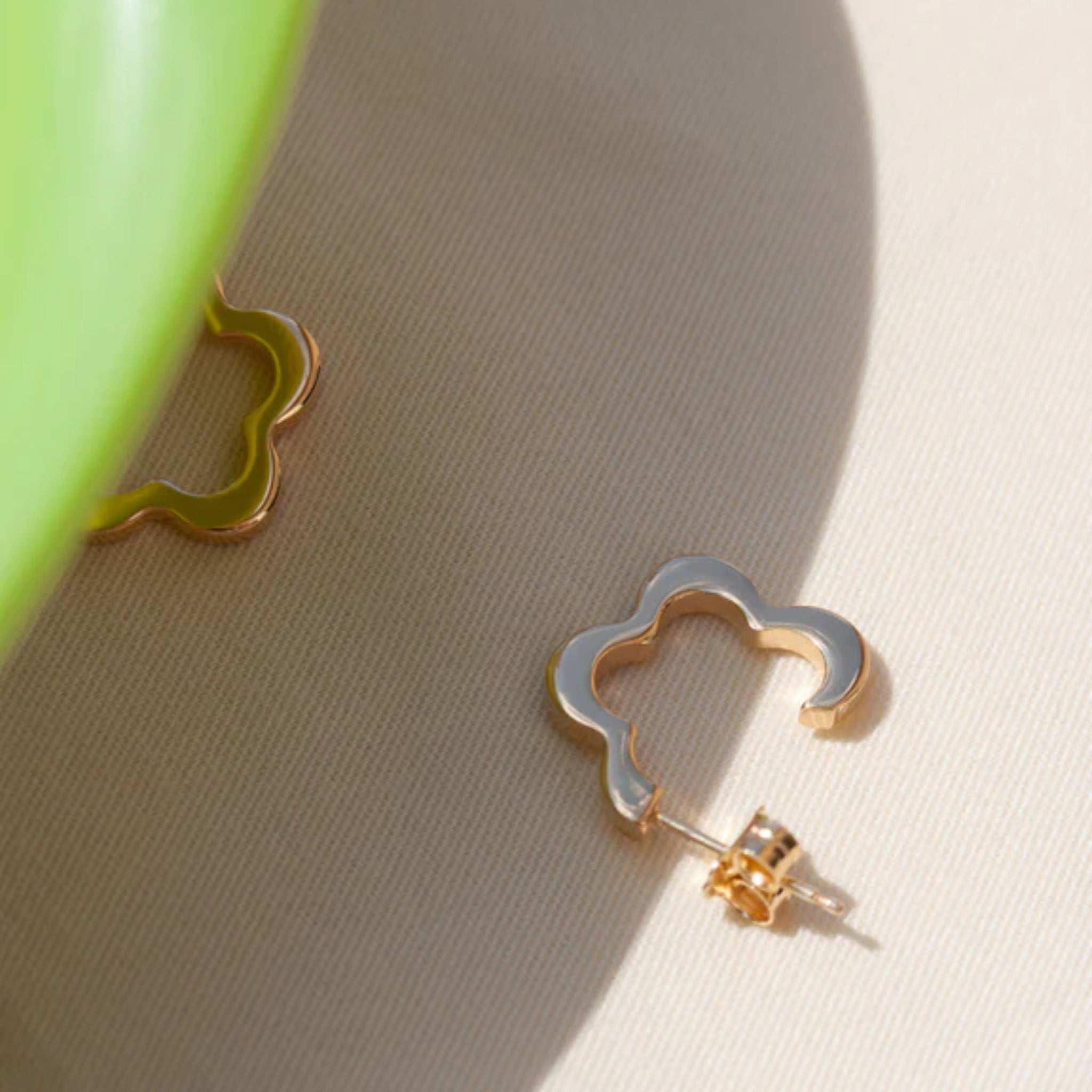 Goldener Ohrring mit geschwungener Form auf beigem Stoff, beleuchtet von Sonnenlicht.