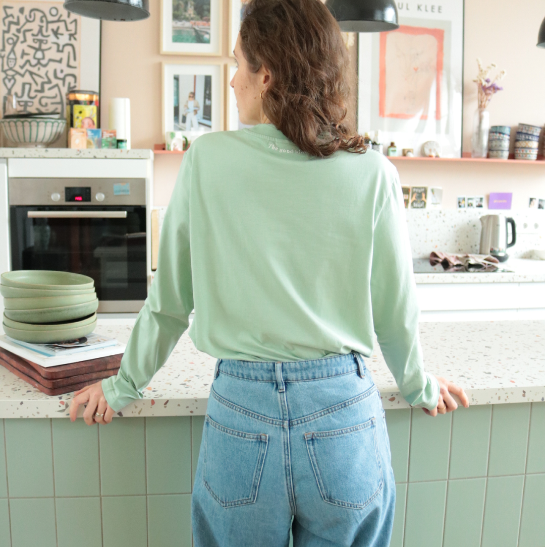 Person steht in einer Küche mit grünen Fliesen, trägt ein grünes Oberteil und Jeans, mehrere Bilder an der Wand.