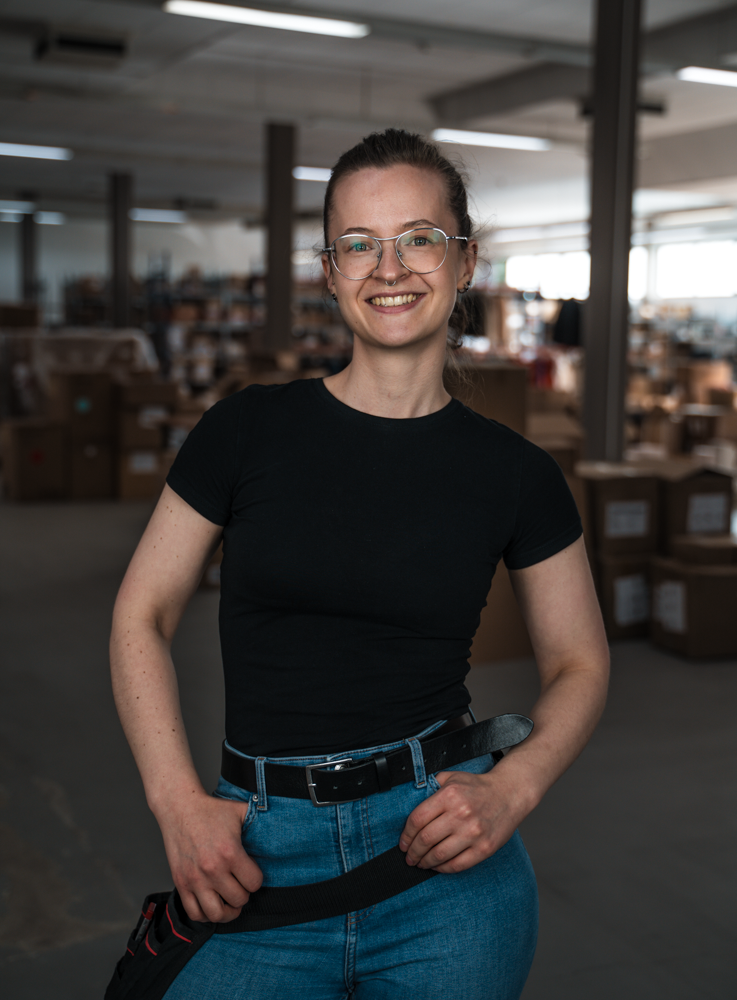 Lächelnde Person mit Brille und dunklem T-Shirt steht in einem Lager voller Kartons.