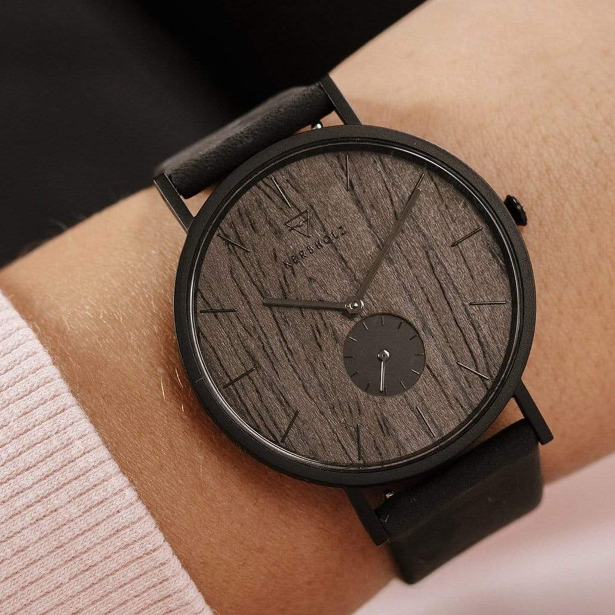 Armbanduhr mit dunkelbraunem Holz-Zifferblatt und schwarzem Lederarmband an einem Handgelenk.