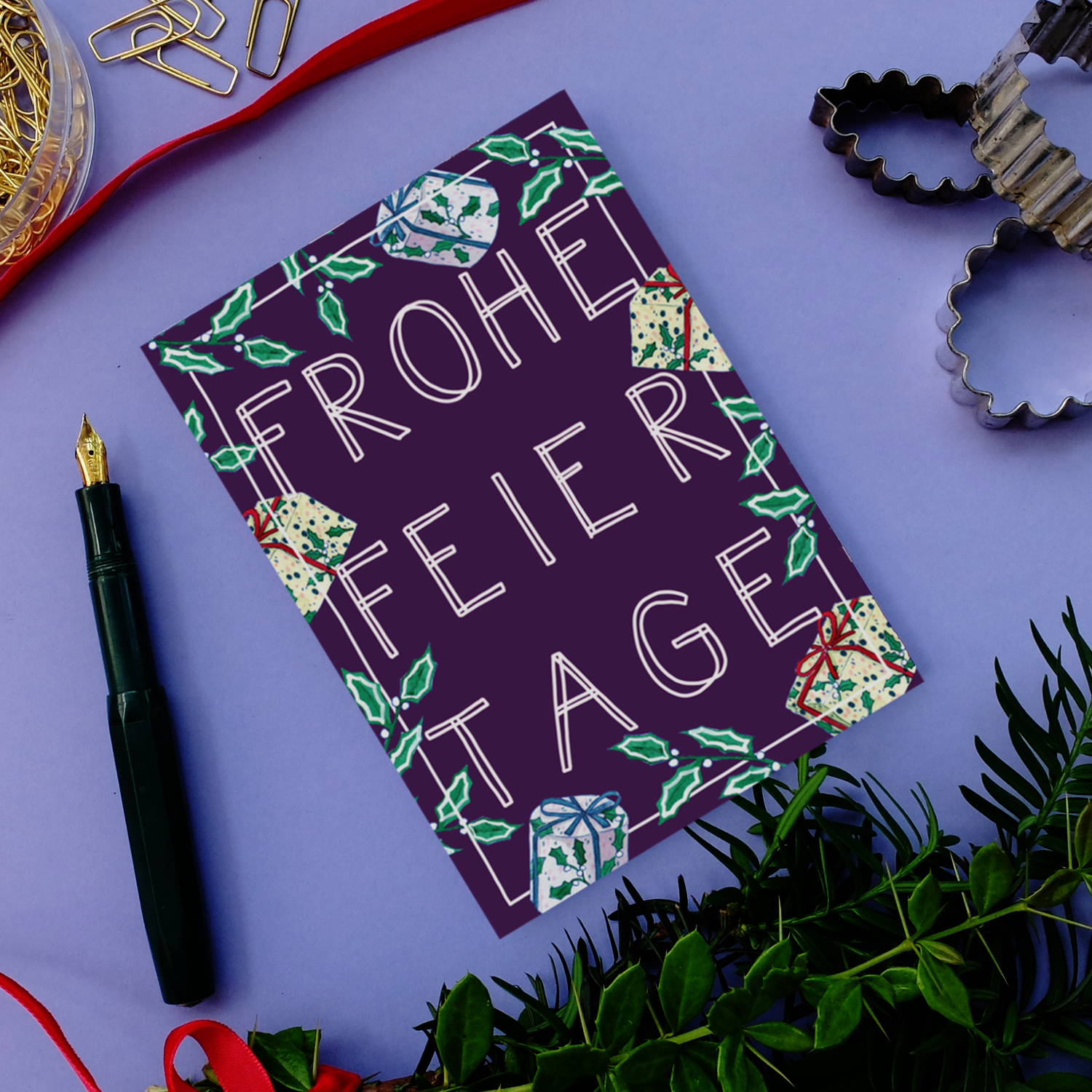 Eine festliche Karte auf lilafarbenem Untergrund mit der Aufschrift "Frohe Feiertage" ist umgeben von Stechpalmen, Geschenken, einem Füllfederhalter, Grünzeug, Büroklammern und Ausstechformen.