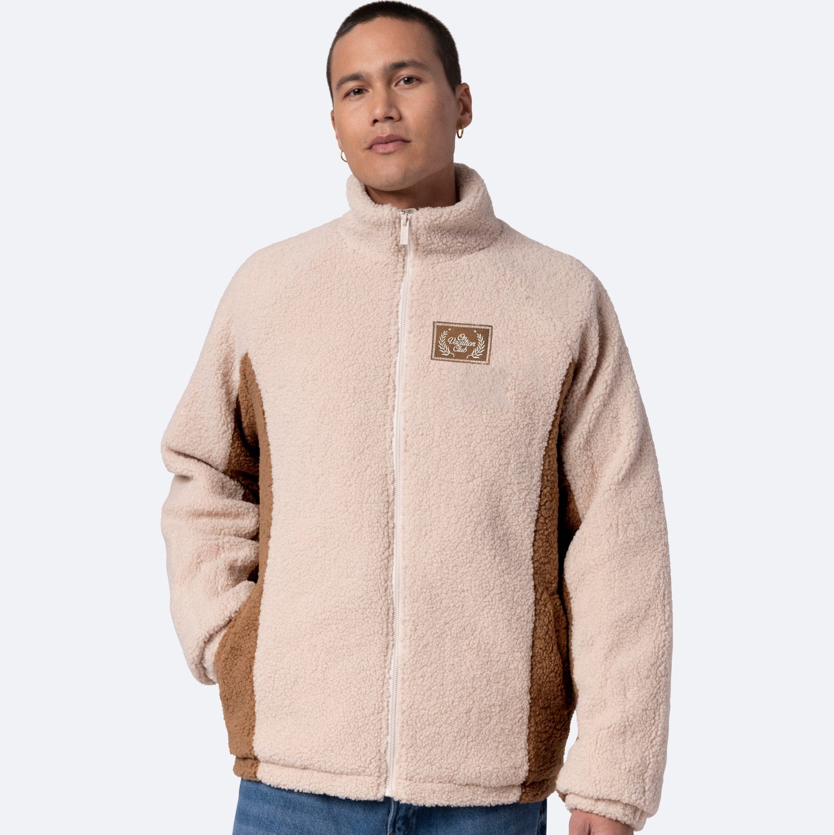 Eine Person, die eine beige und braune Sherpa-Fleecejacke mit Reißverschluss mit hohem Kragen und einem kleinen Aufnäher auf der Brust trägt, gepaart mit blauen Jeans, steht vor einem schlichten hellen Hintergrund.