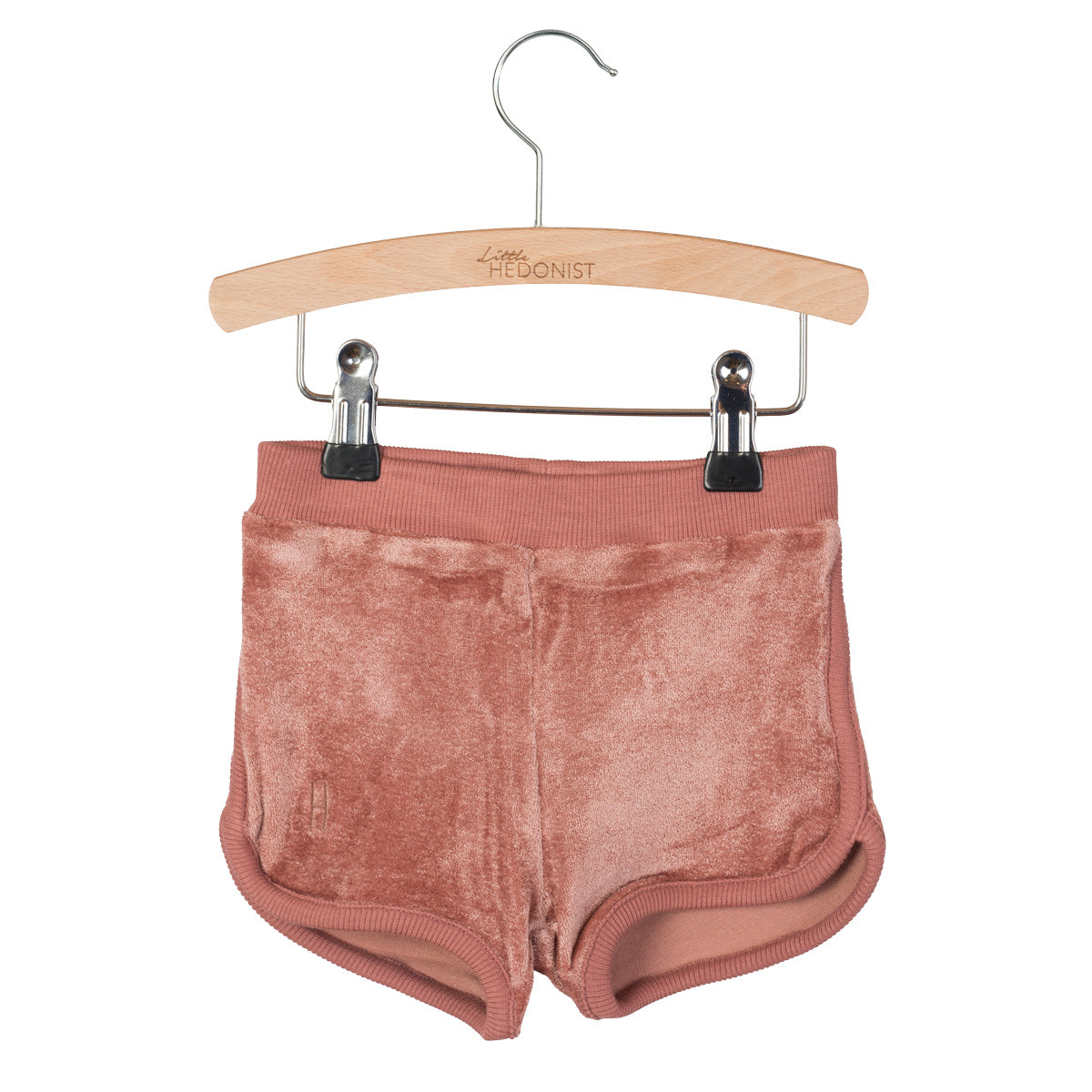Ein Paar weiche, rosafarbene Samtshorts hängt an einem Holzbügel mit Metallklammern. Die Shorts haben einen gerippten Bund und Ränder, und der Bügel ist mit Little HEDONIST beschriftet.