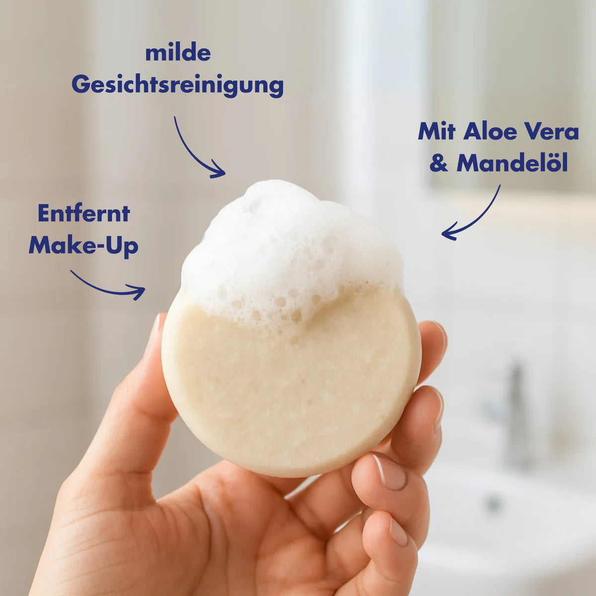 Eine Hand hält einen schaumigen runden Reinigungsstab. Der deutsche Text weist auf seine Eigenschaften hin: milde Gesichtsreinigung, entfernt Make-up und enthält Aloe Vera und Mandelöl. Der Hintergrund ist ein verschwommenes Badezimmer.