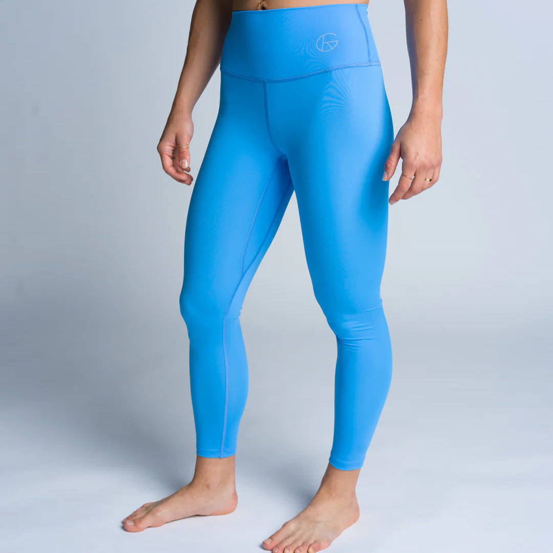 Eine Person, die einen schwarzen Sport-BH und blaue Leggings mit hoher Taille trägt, steht barfuß auf einem hellgrauen Hintergrund, leicht zur Seite gewandt, mit entspannten Armen an der Seite.