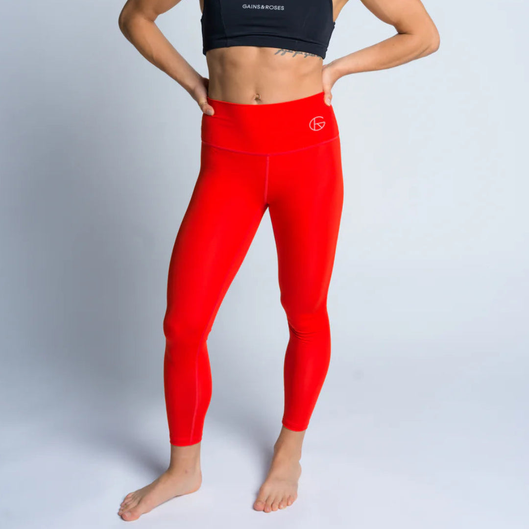 Eine Frau in einem schwarzen Sport-BH und knallroten Leggings steht barfuß vor einem hellen Hintergrund, lächelt, hat die Hände in die Hüften gestemmt und die Haare hochgesteckt.