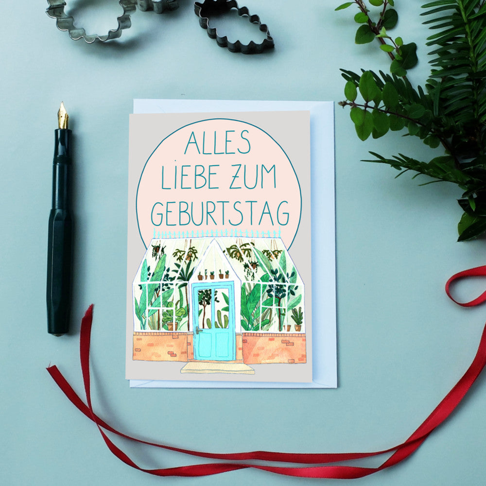 Geburtstagskarte mit der Aufschrift "Alles Liebe zum Geburtstag" und einem illustrierten Gewächshaus auf einem Tisch.