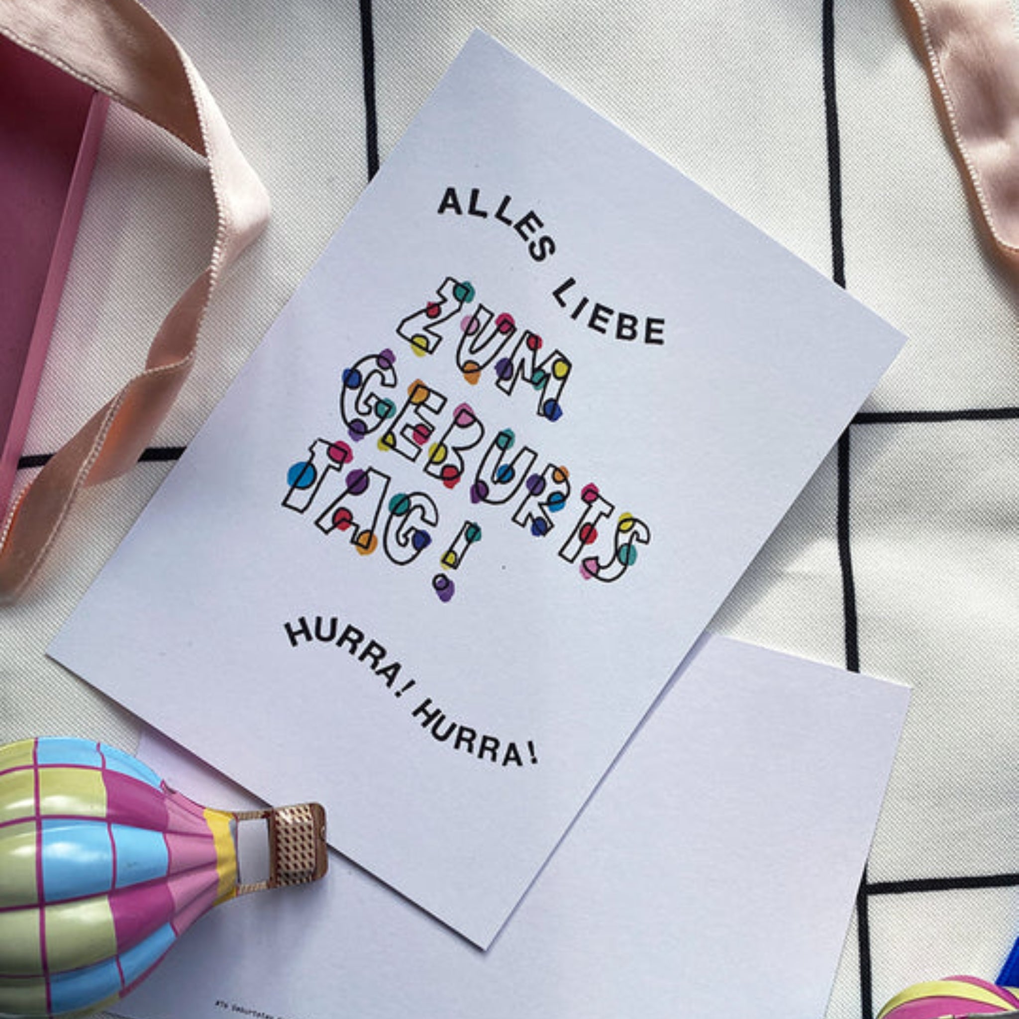 Eine Geburtstagskarte mit der Aufschrift "Alles Liebe zum Geburtstag! Hurra, hurra!" liegt neben einem bunten Spielzeug.