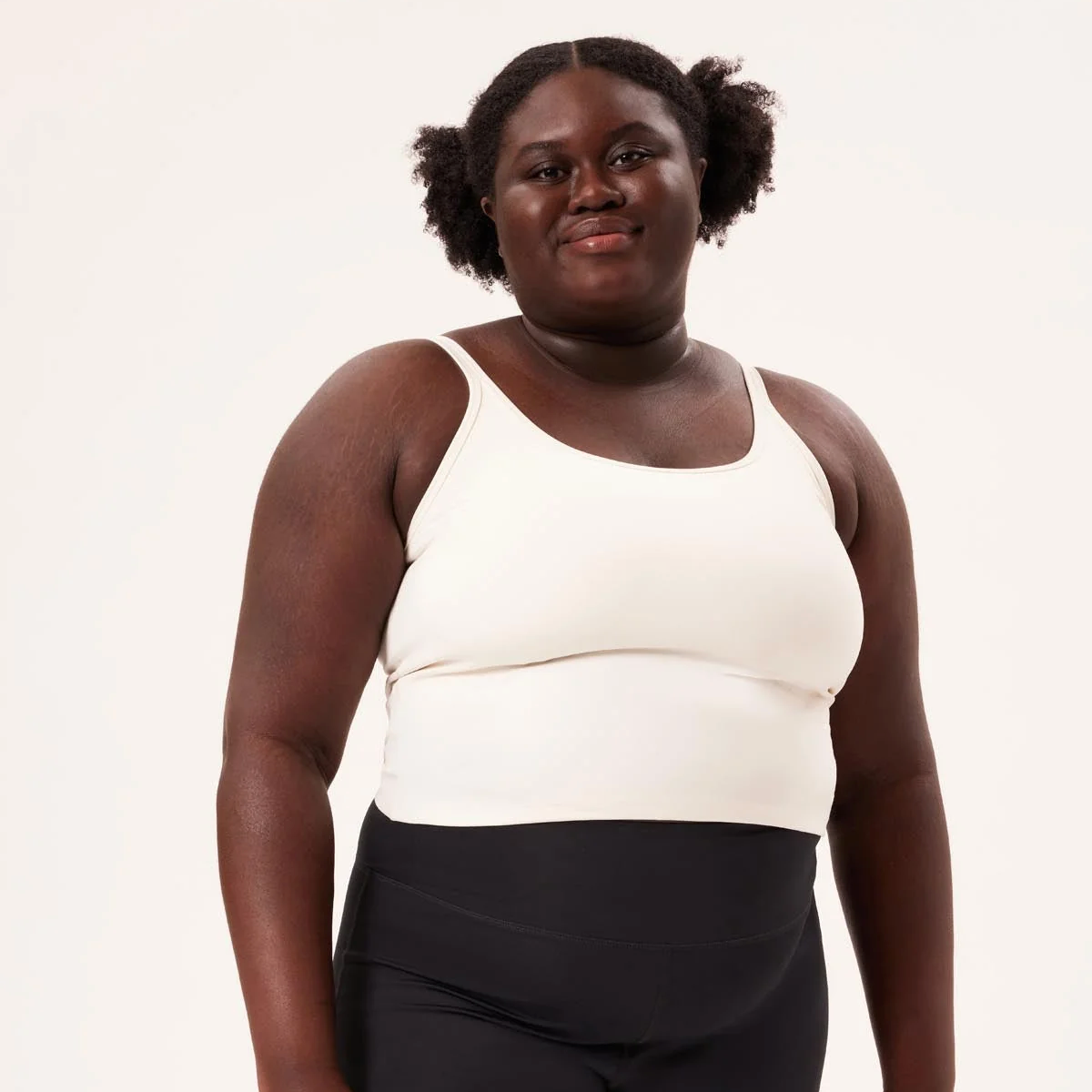 Eine Person mit dunkler Hautfarbe und natürlichem Haar, das in zwei Locken frisiert ist, trägt ein weißes Tanktop und schwarze Leggings, steht vor einem schlichten hellen Hintergrund und lächelt leicht.