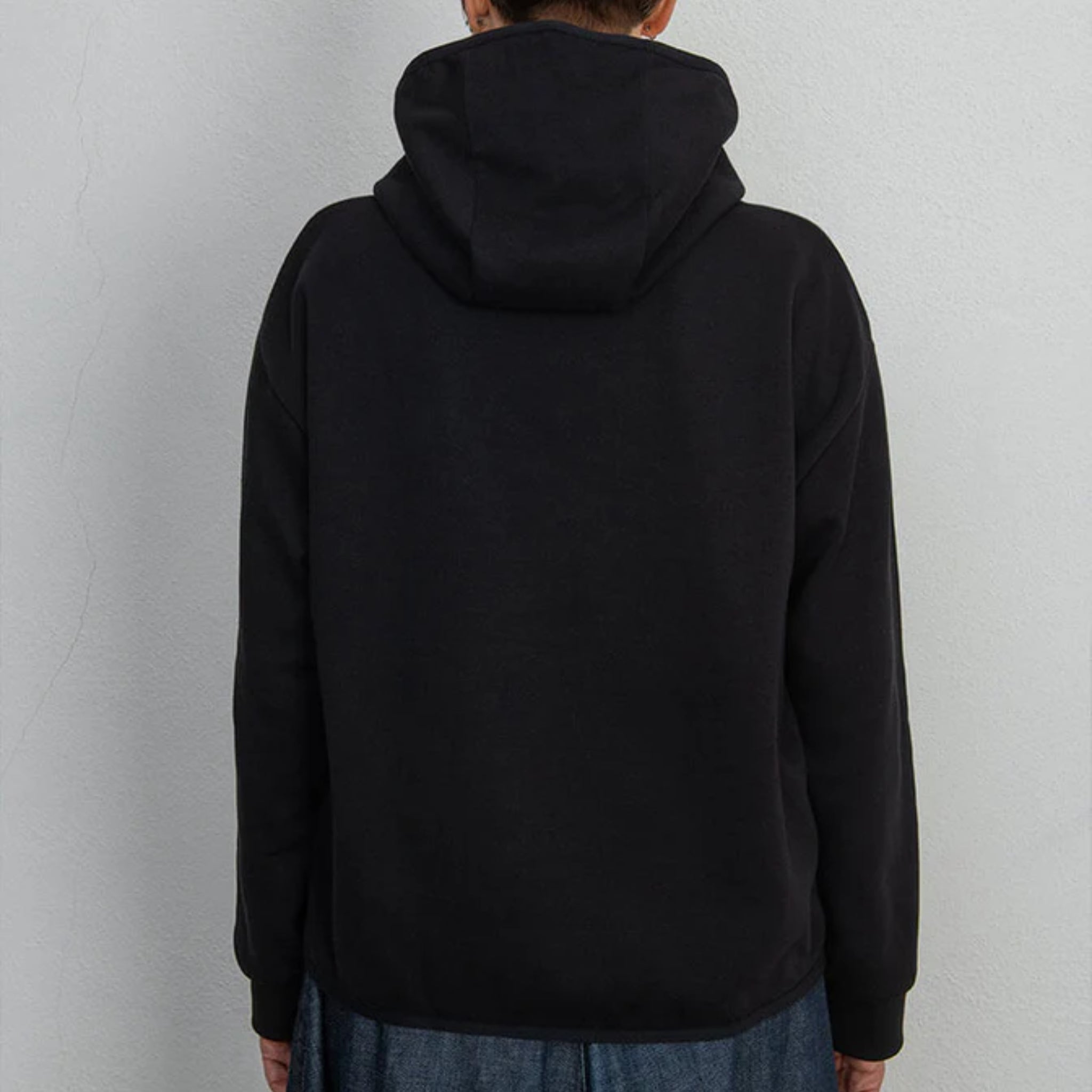 Eine Person, die ein schwarzes Kapuzensweatshirt und eine blaue Jeans trägt, steht vor einer weißen Wand, von hinten gesehen.