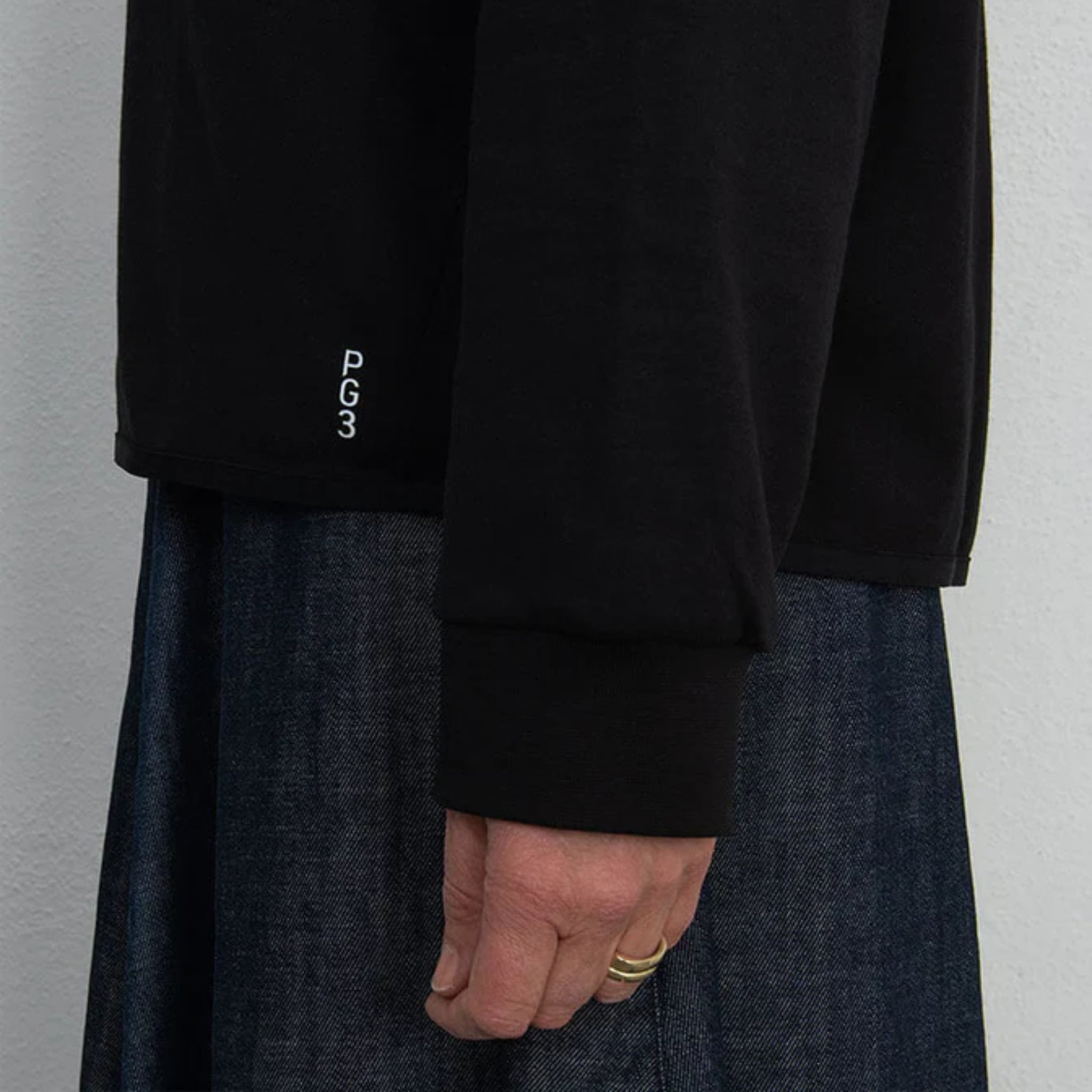 Eine Person, die ein schwarzes langärmeliges Hemd mit der Aufschrift PG 3 am Saum und eine dunkelblaue Jeanshose trägt, steht vor einer schlichten hellen Wand. Nur die Seite ihres Oberkörpers und ihre Hand sind sichtbar.
