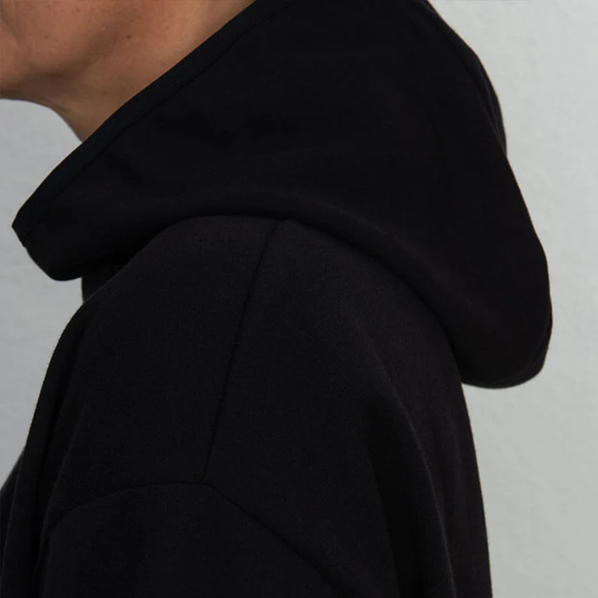 Nahaufnahme einer Person, die einen schwarzen Kapuzenpulli trägt, wobei ein Teil des Gesichts, des Halses und der Schulter vor einem einfarbigen hellen Hintergrund zu sehen ist.