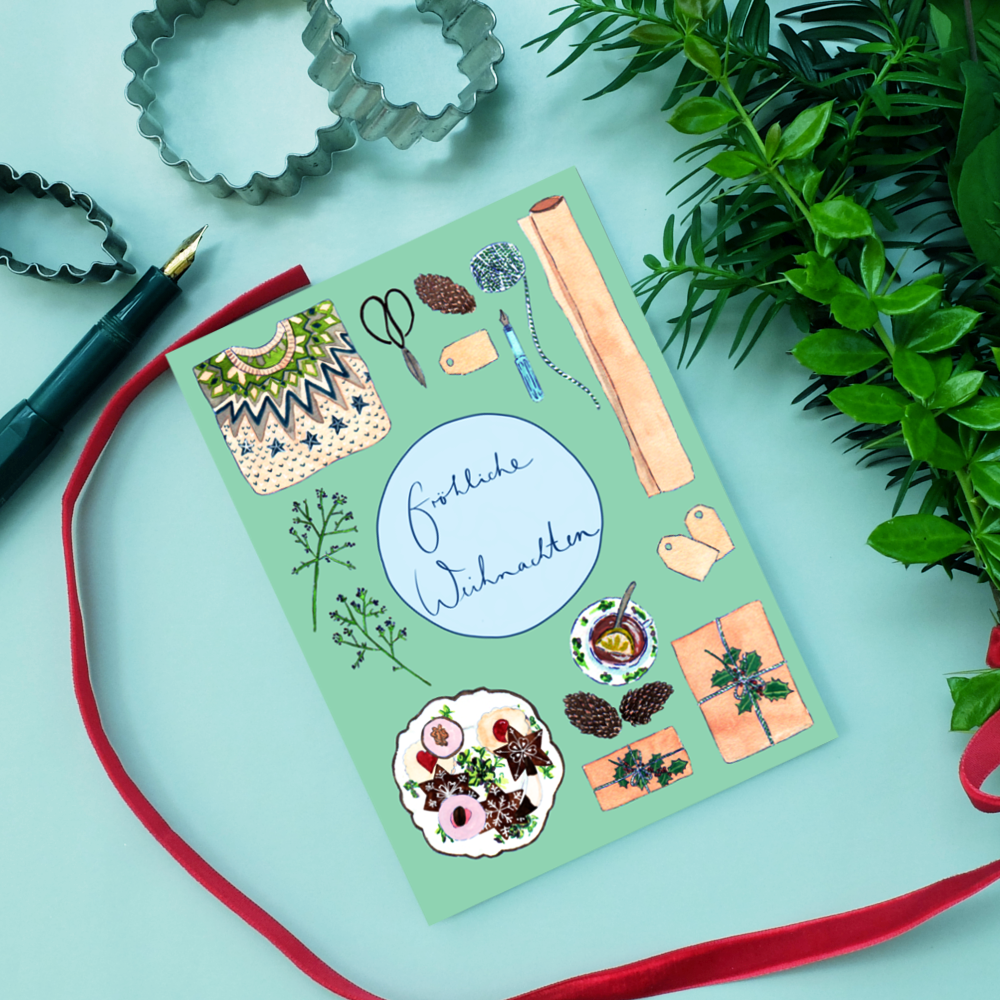 Ein festliches Flat Lay mit einer deutschen Weihnachtskarte mit "Fröhliche Weihnachten", umgeben von illustrierten Keksen, Tannenzapfen, Zweigen, Anhängern, einem roten Band und einem Stift, mit Grünzeug in der Ecke.