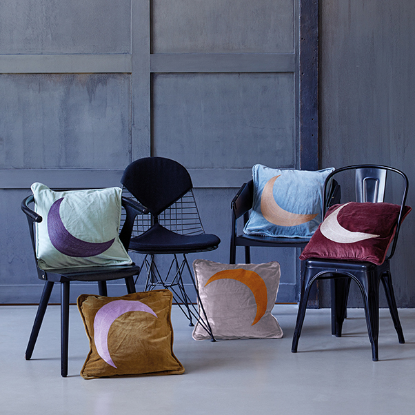 Drei Stühle mit bunten Kissen, die Halbmonde zeigen, stehen vor einer grauen Wand auf hellem Boden.