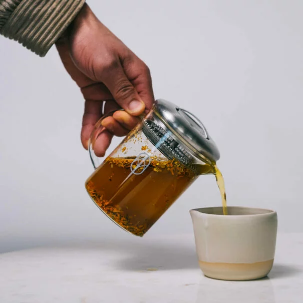 Eine Hand gießt Tee aus einer Glaskanne in eine weiße Tasse auf einem Tisch.