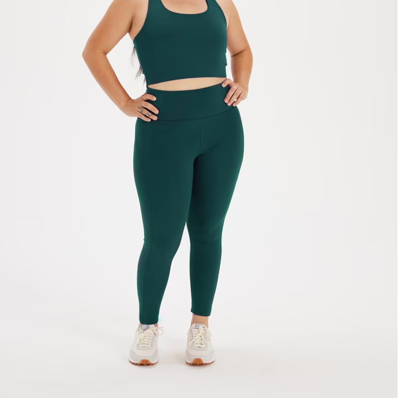 Eine Frau, die einen dunkelgrünen Sport-BH und dazu passende hoch taillierte Leggings trägt, steht mit einer Hand auf der Hüfte. Sie trägt außerdem weiße Turnschuhe. Der Hintergrund ist einfarbig weiß.