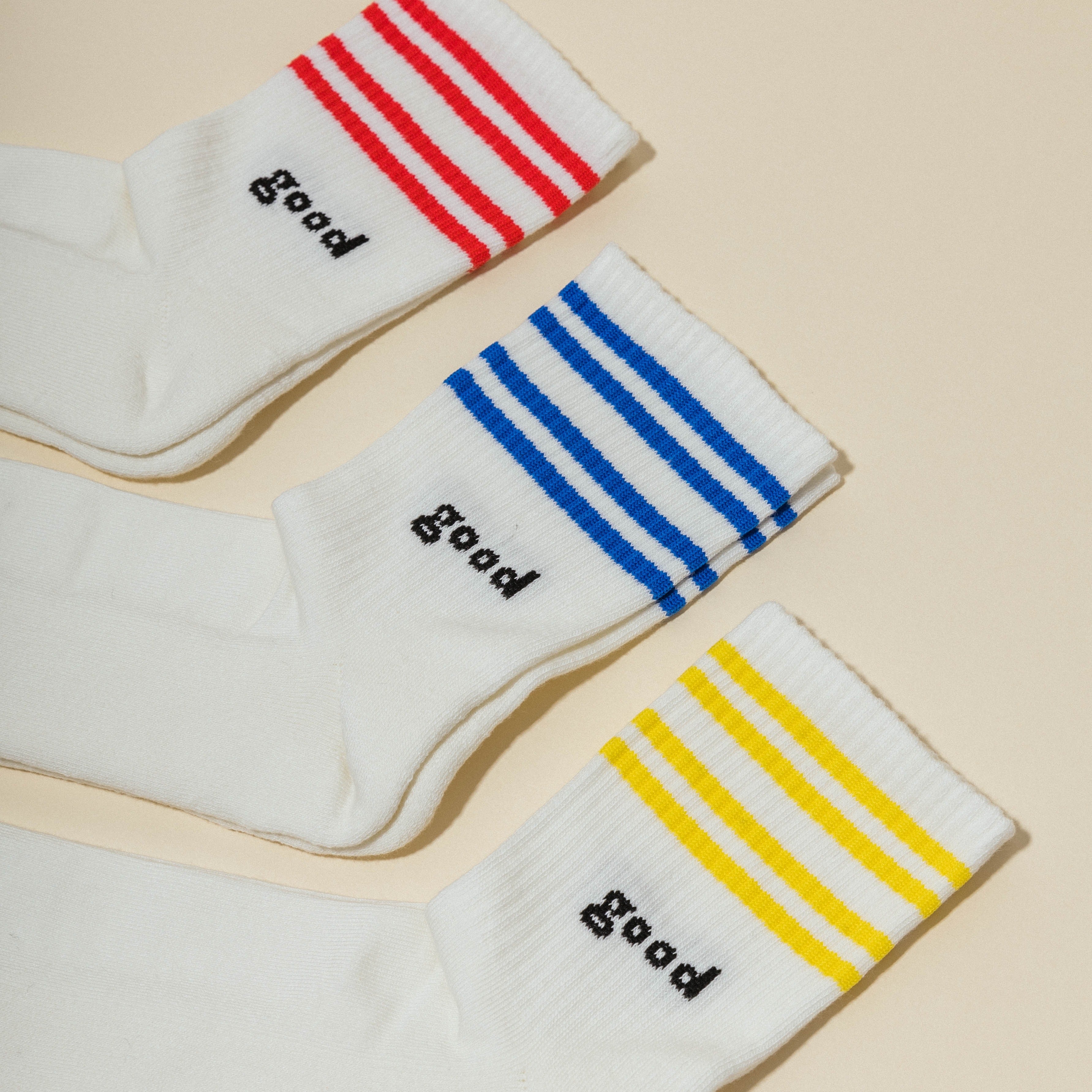 Drei Paar weiße Socken mit Streifen in Rot, Blau und Gelb, jedes mit dem Wort "good".