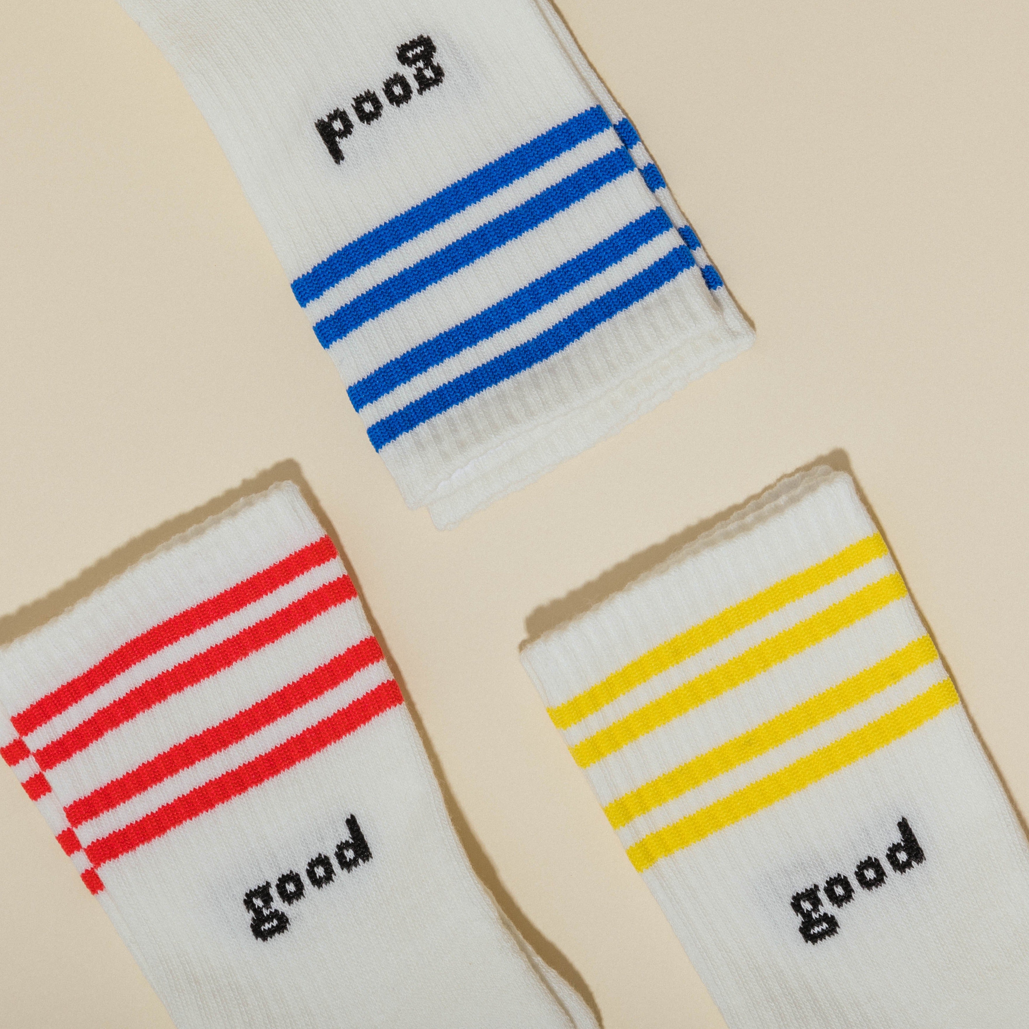 Drei weiße Socken mit bunten Streifen und dem Wort "good" in Blau, Rot und Gelb auf beigem Hintergrund.