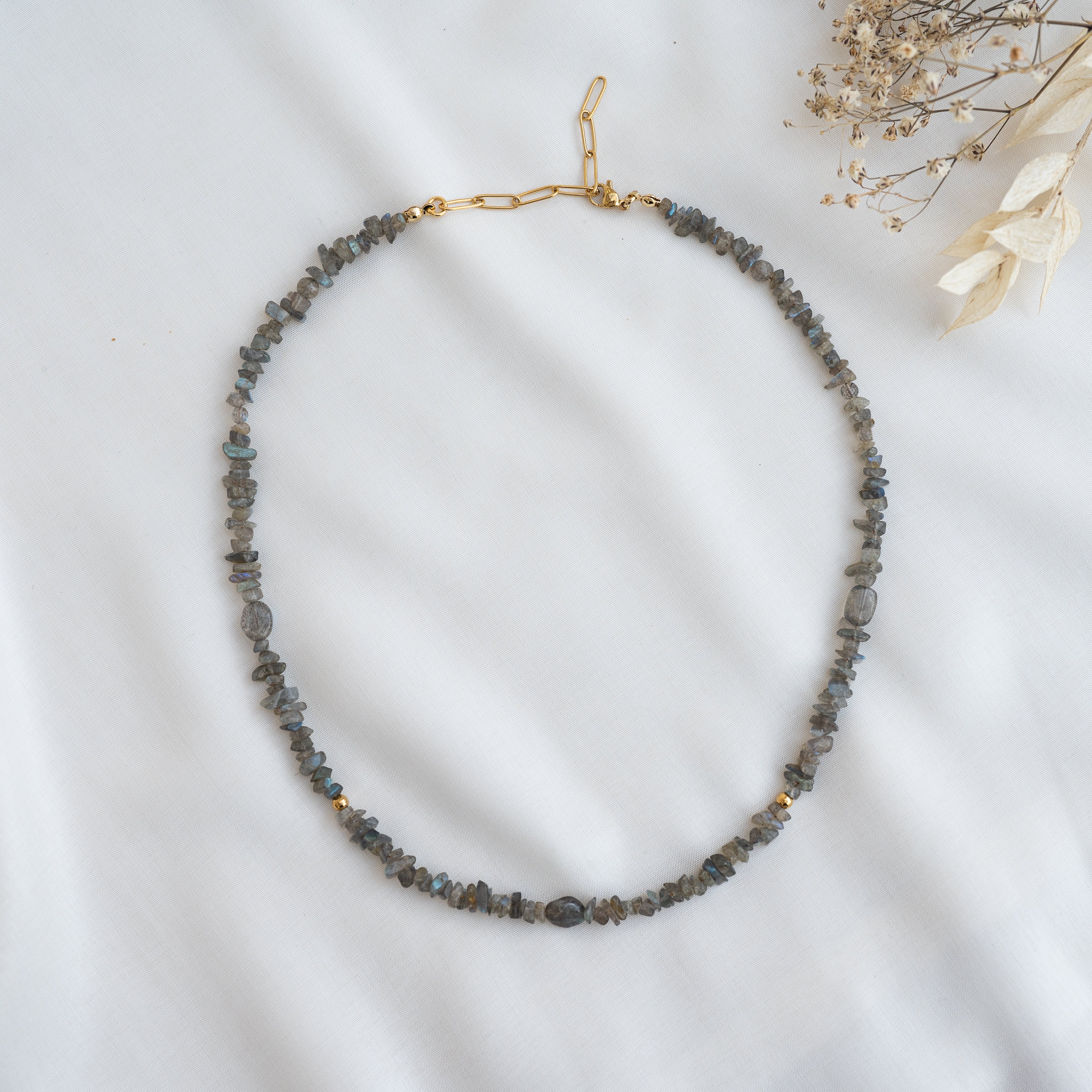 Eine Perlenkette mit unregelmäßigen grauen Steinen und kleinen goldenen Akzenten, kreisförmig auf weißem Stoff mit getrockneten Blumen in der rechten oberen Ecke.