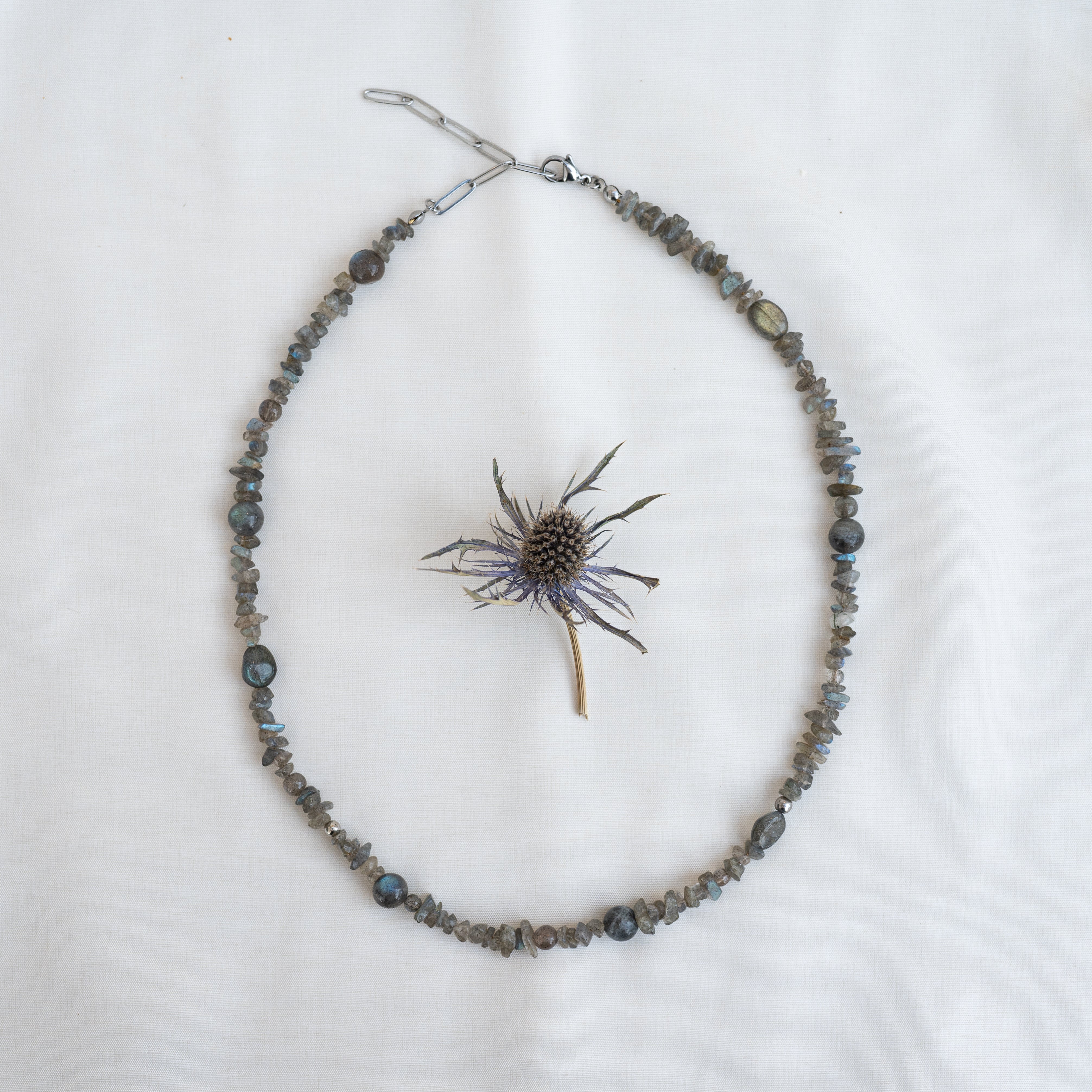 Eine mit Perlen besetzte graue Edelsteinkette, kreisförmig auf einem weißen Tuch angeordnet, mit einer einzelnen getrockneten Distelblüte in der Mitte der Kette.