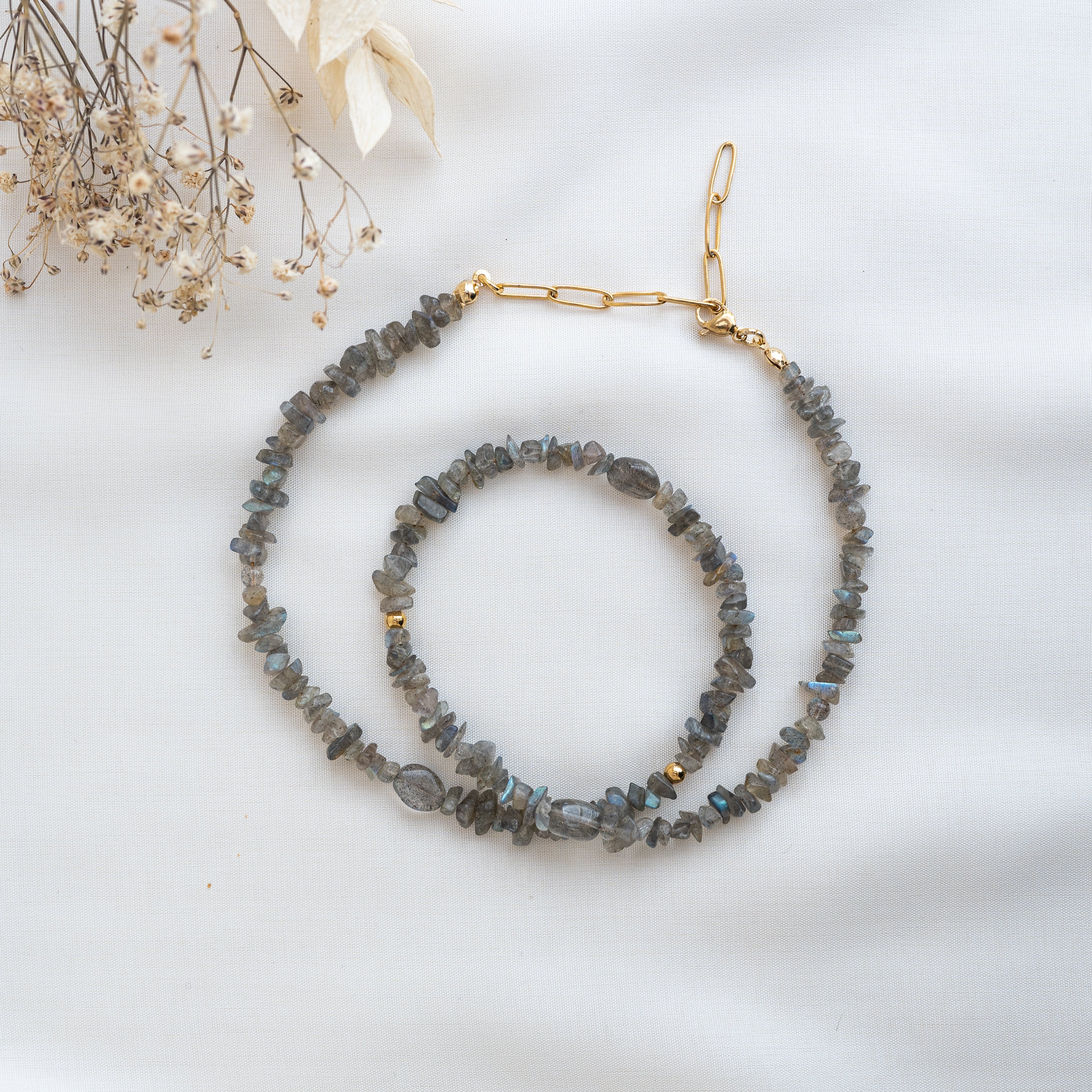 Eine Labradorit-Edelsteinkette und ein Armband mit unregelmäßig geformten Steinen und goldenen Akzenten sind auf einem leichten Stoffhintergrund neben getrockneten Blumen ausgestellt.