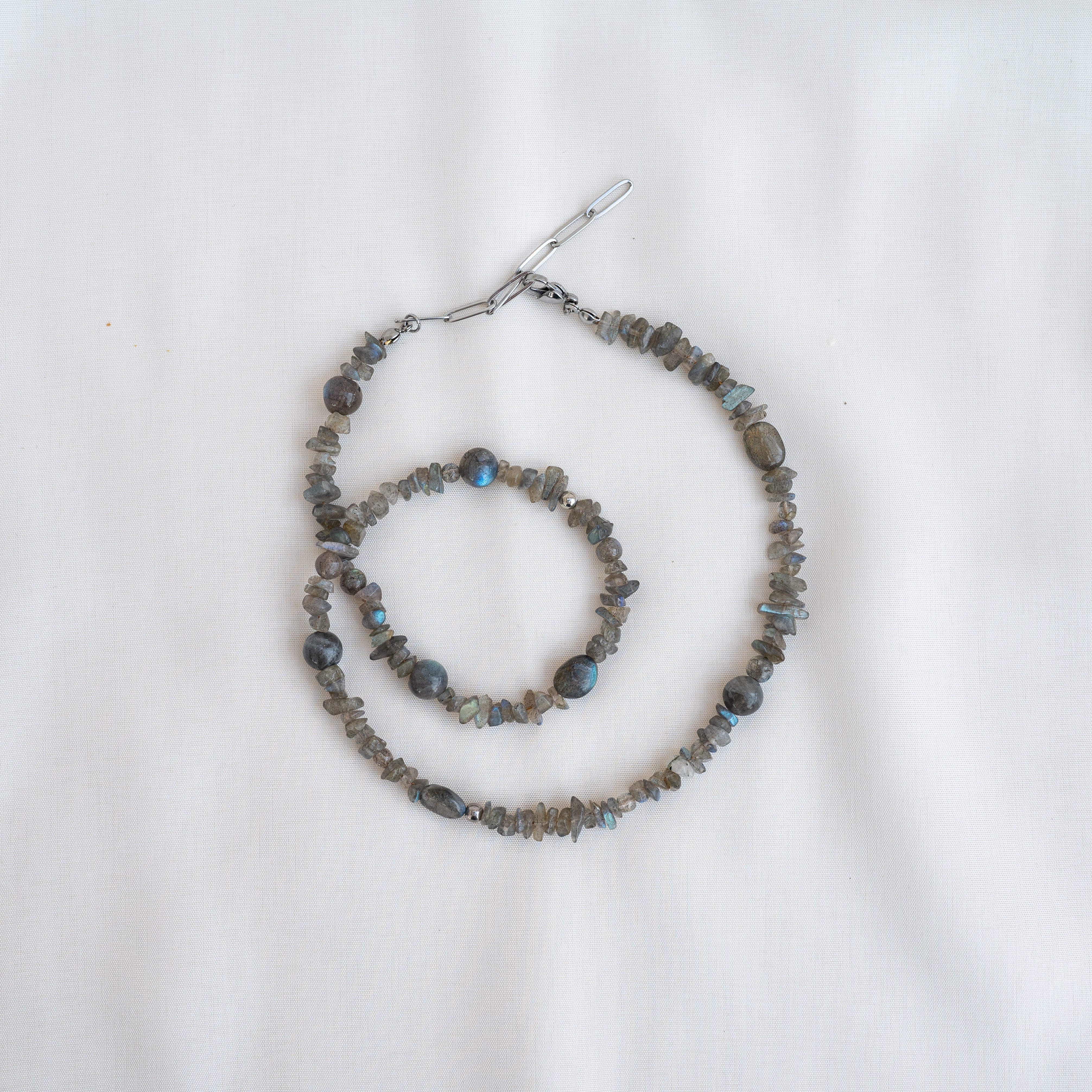 Eine graue Perlenkette und ein passendes Armband mit unregelmäßig geformten Steinen auf einem schlichten weißen Stoffhintergrund.