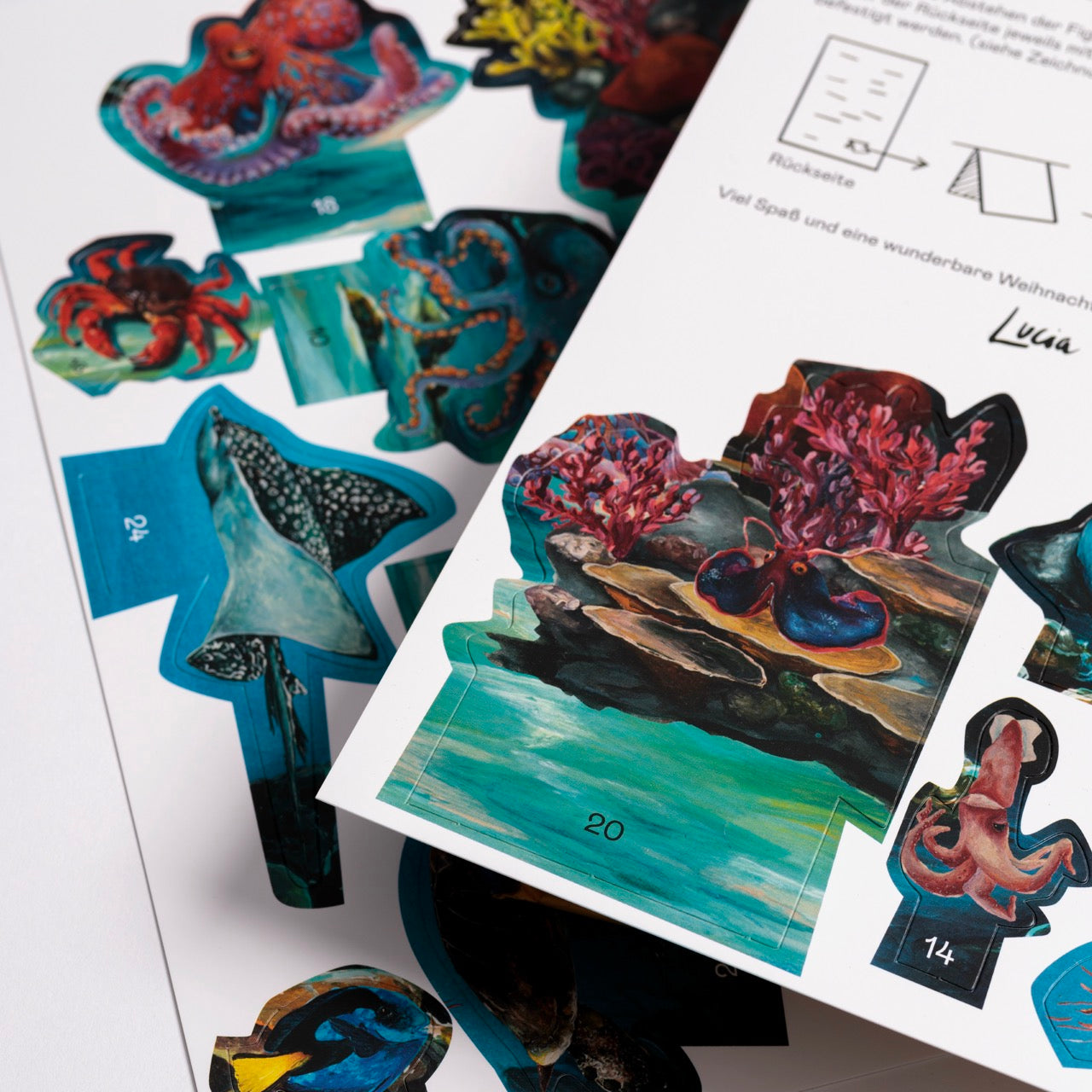 Bögen mit bunten ausgeschnittenen Papierformen, die Meerestiere wie Kraken und einen Mantarochen zeigen, mit deutschen Anweisungen, die teilweise auf dem weißen Papier über den Ausschnitten sichtbar sind.