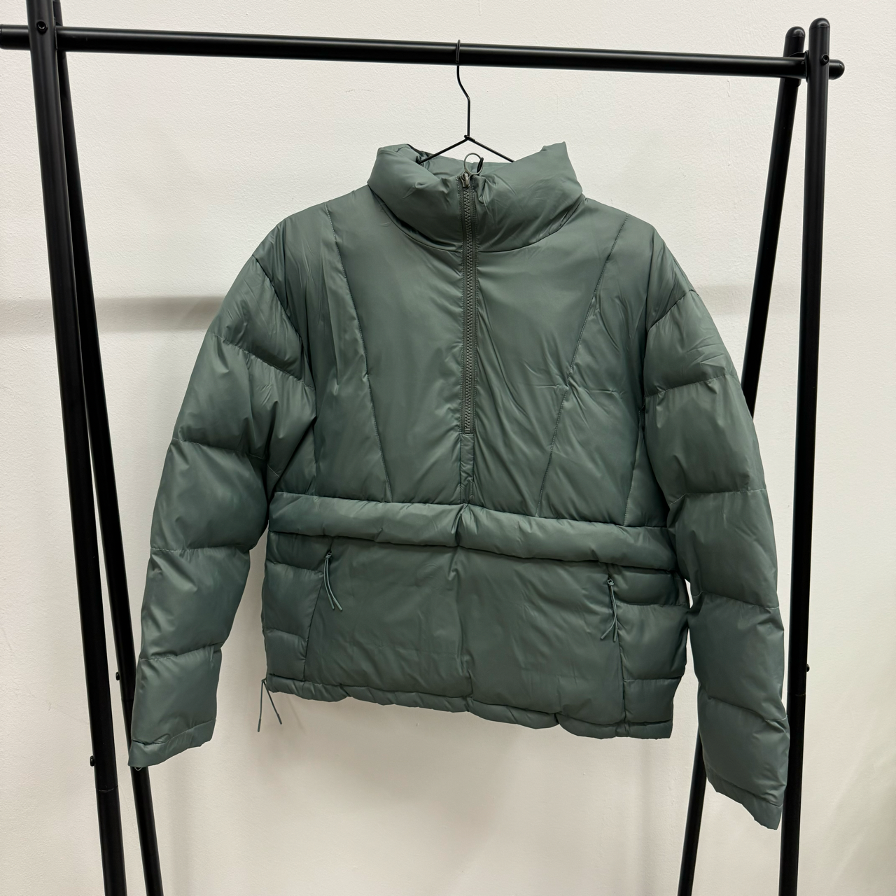 Grüne, gesteppte Daunenjacke mit hohem Kragen und Reißverschluss hängt an einem schwarzen Ständer vor weißer Wand.
