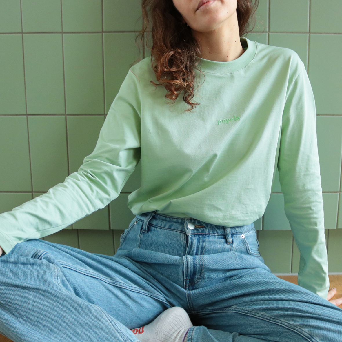 Person in mintgrünem Langarmshirt und Jeans sitzt auf einem Boden vor grünen Fliesen, leicht nach vorne gebeugt.