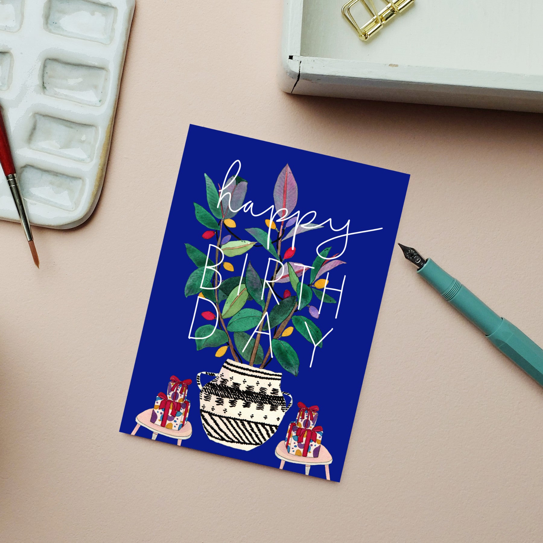 Eine Geburtstagskarte mit einer Pflanze in einer gemusterten Vase, bunten Blättern und der Aufschrift "Happy Birthday" in Weiß. Die Karte liegt auf einer rosafarbenen Fläche neben einem Füllfederhalter, einer Farbpalette und einer weißen Schachtel mit goldenen Klammern.