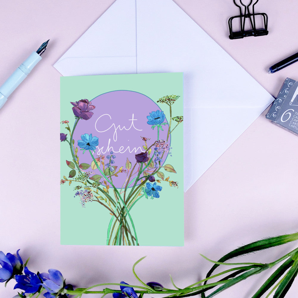 Grußkarte mit Blumen und dem Schriftzug "Gut sben" auf lila Hintergrund, daneben ein Umschlag und Stift.